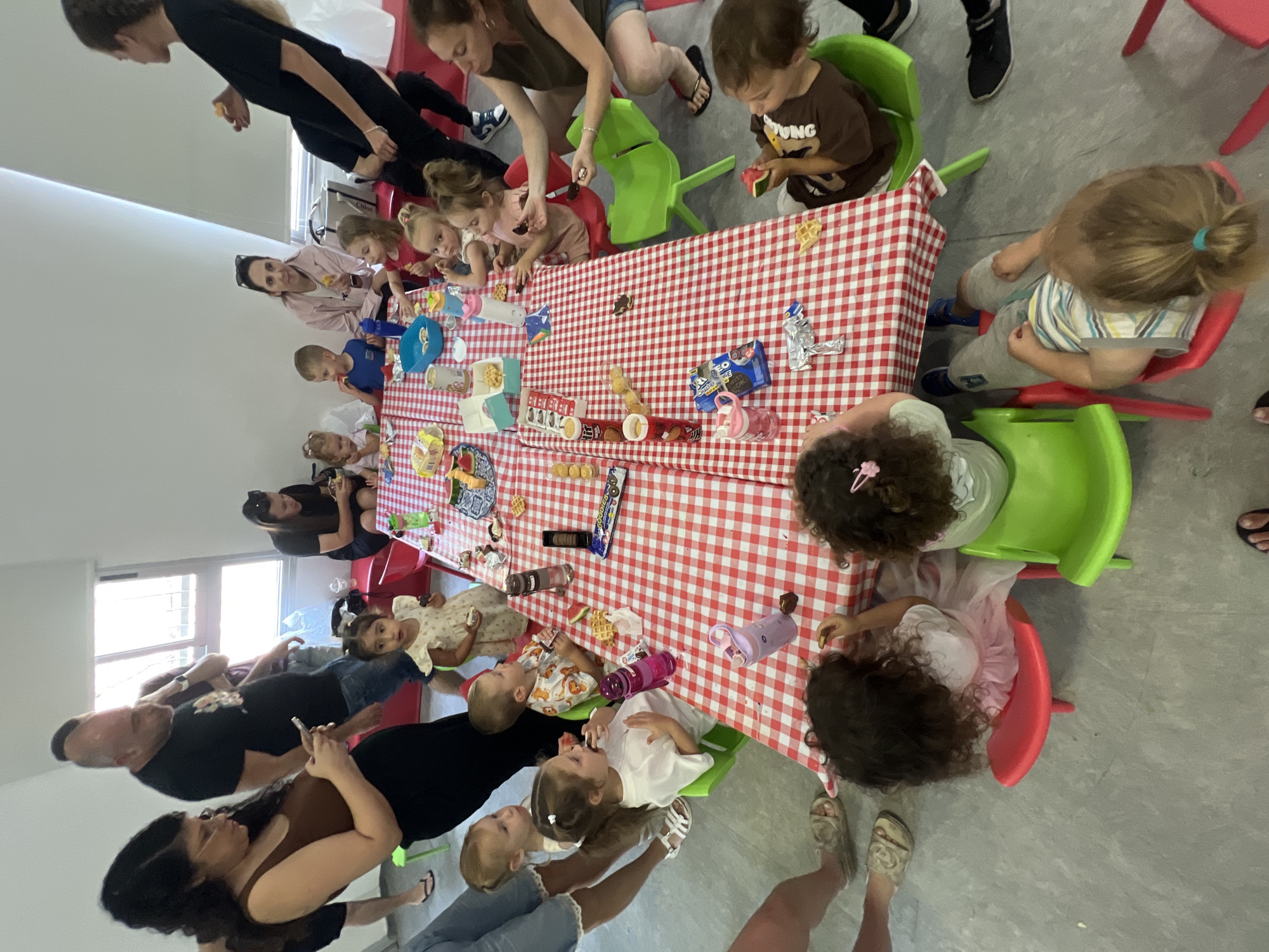 This media file captures a gathering of adults and children around a table covered with a red and white checkered tablecloth. The subjects are primarily young children seated at small, brightly colored plastic chairs, interacting with snacks and drinks laid out on the table. Several adults are also present, observing and possibly assisting the children.
The scene appears to be an indoor setting, likely a community center or a home, given the plain gray floor and the presence of windows in the background. The overall atmosphere seems relaxed and informal, suggesting a party or a shared meal. The children are engaged with their food, some are eating, while others are looking at the items on the table.
Notable details include various food items and beverages spread across the table, such as waffles, candies, and what appear to be juice boxes or water bottles. Some of the children are holding these items, indicating they are actively participating in the event. The clothing of the subjects is casual, consistent with a daytime gathering.
There is no visible text in the image that provides specific information about the event, time of day, or weather. However, the bright lighting suggests it is daytime. The emotions conveyed are generally positive, with children appearing engaged and adults seemingly content. The context provided indicates the location is Kiryat Bialik, Israel.