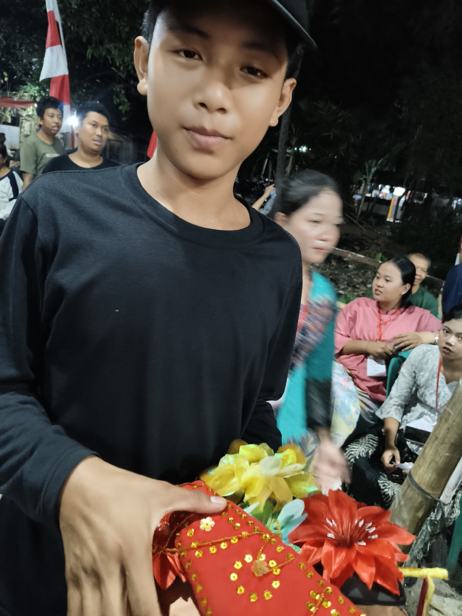 The image depicts a close-up of a young male wearing a black long-sleeved shirt and a black cap. He is holding a small red gift box adorned with gold sequins and artificial flowers in red and yellow. The background is dimly lit and suggests an outdoor evening setting. Several other individuals are visible in the background, blurred and out of focus. Two people are wearing traditional Indonesian clothing. A red and white Indonesian flag is also visible in the background. The overall scene appears to be a gathering or event. Based on the presence of the Indonesian flag and the overall context, the location is likely South Tangerang, Indonesia, possibly during a local festival or celebration.