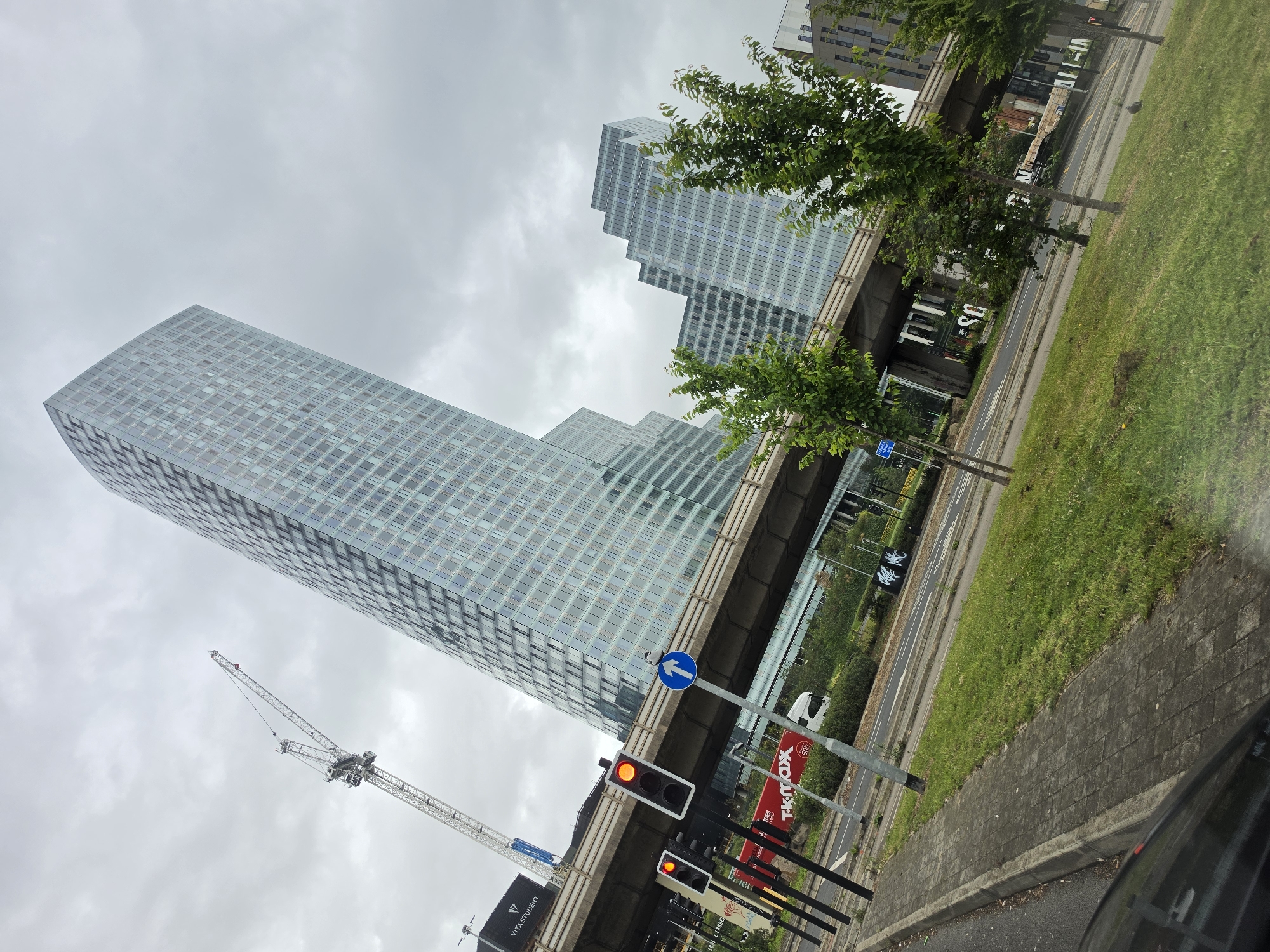 The image captures an overcast urban scene in Manchester, United Kingdom, specifically depicting the area around **Deansgate Square** and **Spinningfields**. The perspective is from a street level, looking upwards at modern architecture.
A dominant feature is a tall, curved, glass-clad skyscraper, identified as **One Spinningfields**, situated prominently on the left-center, its reflective surface mirroring the grey sky. To its right, a substantial elevated concrete flyover or motorway (likely part of the Mancunian Way) traverses horizontally across the middle ground. Beyond this elevated structure, several other contemporary glass-fronted buildings with a stepped design, characteristic of the **Deansgate Square** development, are visible under the overcast sky.
On the far left, a tall white lattice construction crane extends upwards, signifying ongoing development. Below the crane, a dark building with "VITA STUDENT" signage is partially visible. In the immediate foreground on the left, three sets of traffic lights display red (or orange-red) signals, next to a blue circular road sign indicating a straight-ahead direction.
Beneath the elevated road, at ground level, a multi-lane asphalt road is visible. A red vehicle, possibly a truck or bus, bearing "T.K.maxx" and "60%" advertising, is partially seen on the road, alongside a white van. Two indistinct human figures are walking on the road or pavement beneath the flyover. The foreground includes a grey block-paved sidewalk, strips of green grass, and several young, leafy green trees. The bottom-left corner shows the dark roof and interior elements of the vehicle from which the photograph was taken.