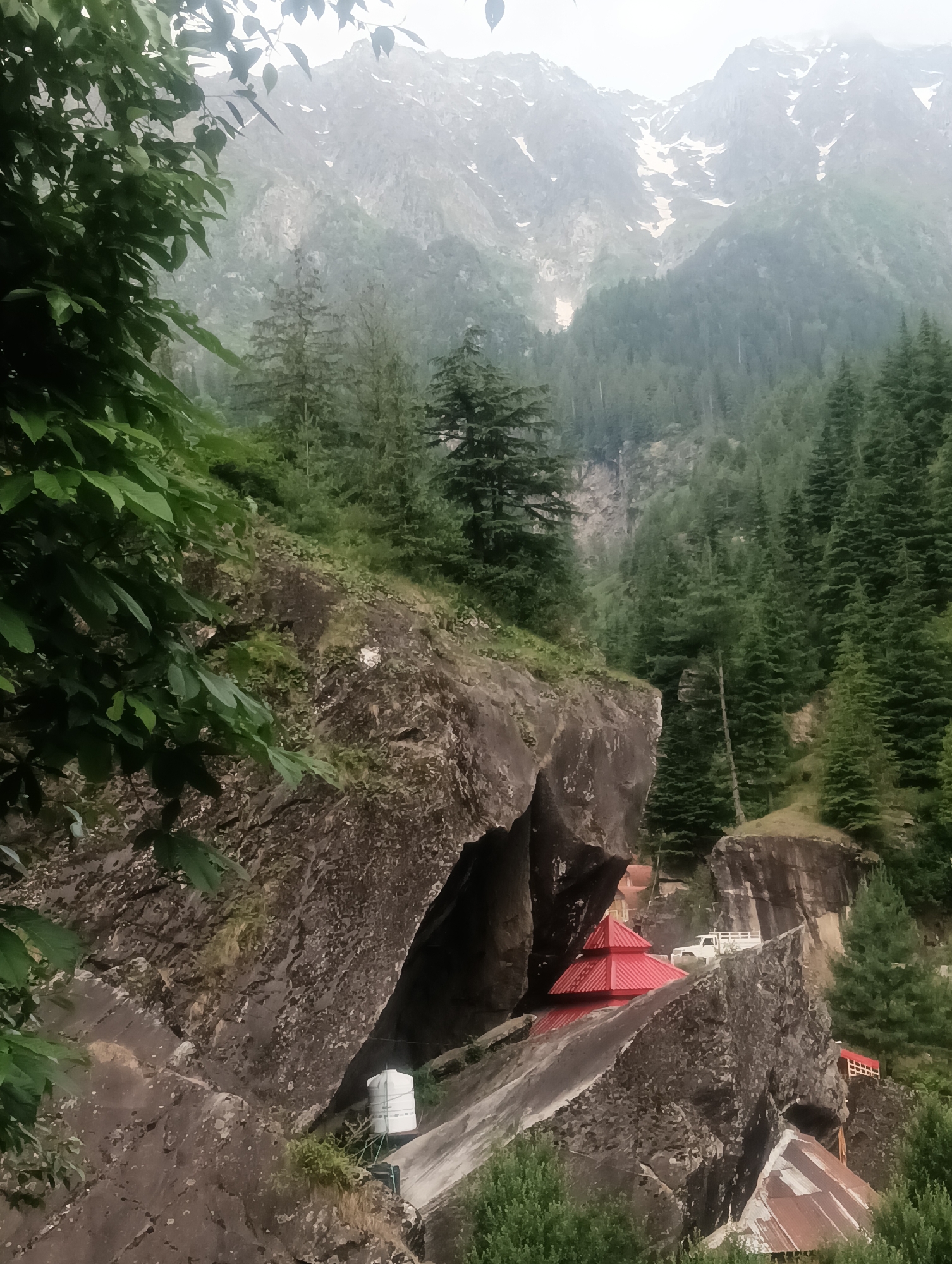 The image depicts a mountainous landscape in Shanag, India, characterized by steep, rocky inclines covered in dense coniferous forests. The foreground features large, dark rock formations, partially obscured by the lush green foliage of trees and bushes. To the right, nestled within the rocks, is a structure with a distinctive red, multi-tiered roof, possibly a temple or a building with traditional architecture. A white truck is parked nearby, suggesting human presence and activity. Further in the background, towering snow-capped mountains rise majestically, partially shrouded in mist, emphasizing the remote and rugged terrain. The overall atmosphere is one of natural grandeur and tranquility.