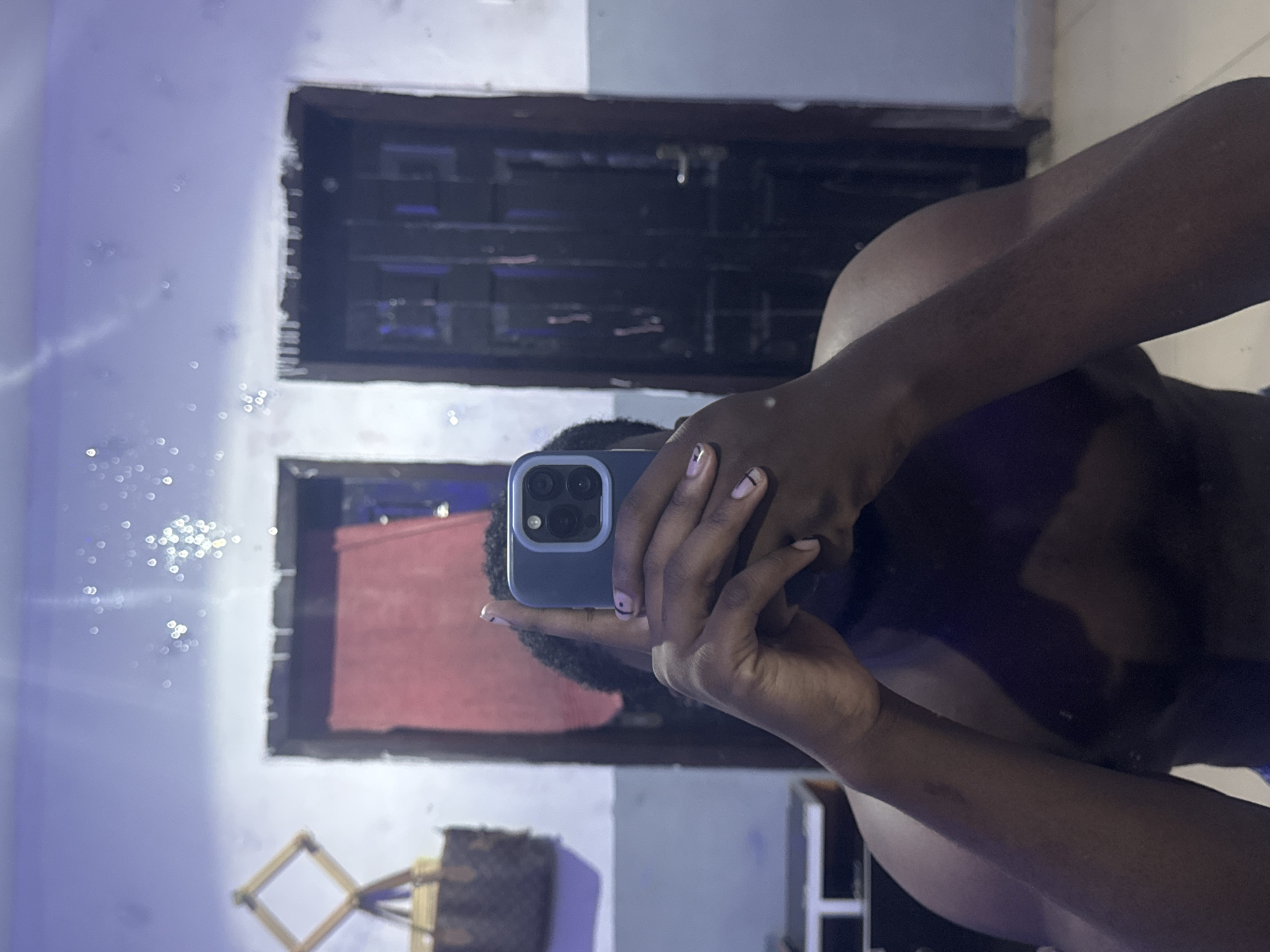 A mirror selfie captures a person holding a smartphone in an indoor setting. The subject, with dark skin, is visible from the mid-torso up on the right side of the frame, holding a blue/gray smartphone, which appears to be an iPhone model, featuring multiple camera lenses. The person's fingernails are painted a light pink hue with small black line accents. They are wearing a dark top or garment.
The background, which is the reflection of the room, features a light-colored wall, possibly white or very pale blue/gray, showing some texture and subtle variations in tone. On the left side of the reflection, numerous small, bright, circular bokeh lights are scattered, suggesting reflections from ambient light sources or decorative string lights.
Two dark, rectangular frames, likely made of wood and resembling door or window frames, are prominent in the background. The upper frame reveals a dark, indistinct area behind it, while the lower frame shows a reddish object, possibly a piece of fabric or furniture. Below the lower frame, towards the bottom left, a wooden, diamond-shaped decorative item or stand is partially visible, next to a brown patterned bag.
The overall lighting appears dim, suggesting an indoor environment, possibly in the evening or night. No discernible text is visible within the image. The scene is set indoors in Gbongan, Nigeria.