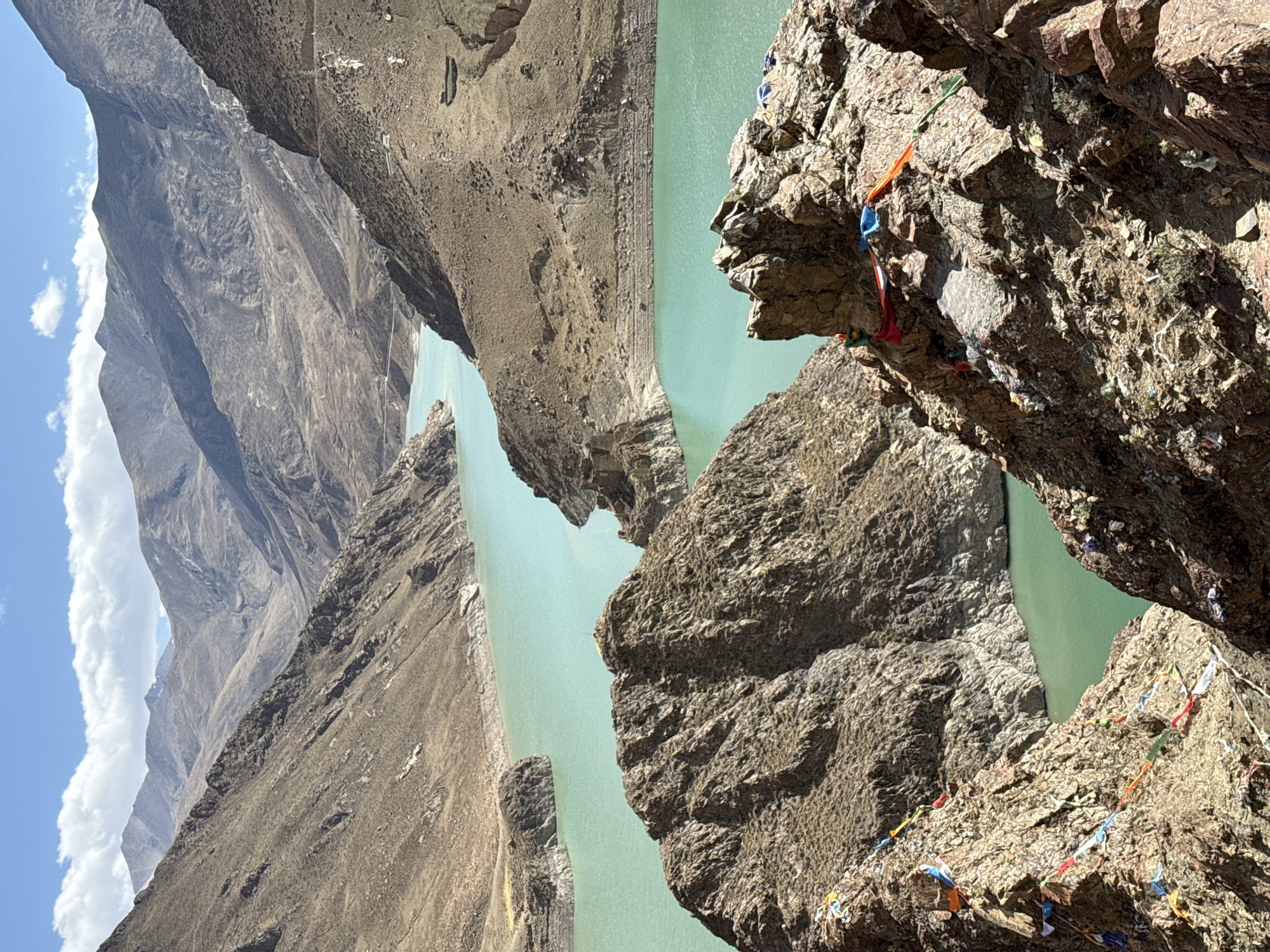 The image depicts a high-altitude landscape in Gyantse County, Shigatse, Tibet Autonomous Region, China, featuring a turquoise lake nestled between steep, rocky mountains. The foreground reveals a rugged rock face adorned with colorful Tibetan prayer flags, suggesting a site of spiritual significance or pilgrimage. The lake, seemingly dammed or contained by the terrain, reflects the sky. Mountains rise in the background under a partly cloudy sky, indicating a sunny day. The scene suggests a remote, mountainous environment with cultural and natural elements coexisting.