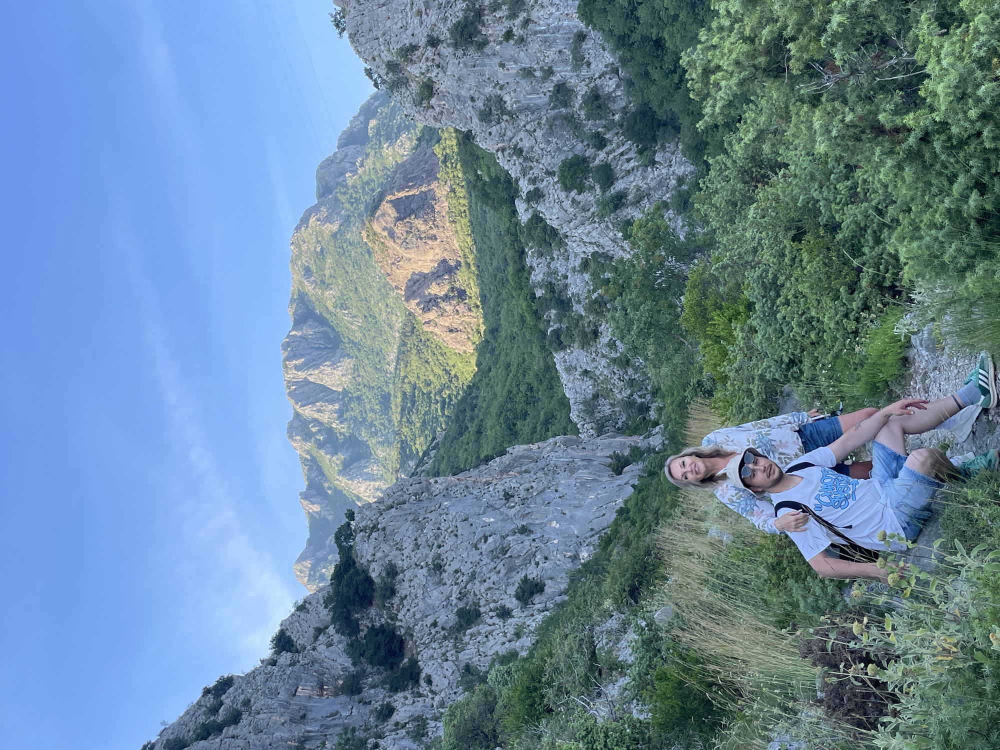 The image, correctly oriented, features two individuals, a man and a woman, seated on a rugged, vegetated mountain slope in Zupci, Montenegro. They are positioned in the foreground, facing the viewer, against a dramatic backdrop of sprawling mountains under a clear blue sky.
The woman, with blonde hair, is wearing a floral long-sleeved top and denim shorts, paired with green and white sneakers. She displays a faint, contented smile. The man, seated slightly in front of her, is wearing a white t-shirt with an abstract blue graphic, denim shorts, a light-colored bucket hat, and sunglasses. He also wears green and white sneakers and has a black backpack. The woman's arm is gently around the man's back, while his arm rests around her waist, suggesting a close relationship as they pose for the photo.
Behind the couple, the immediate terrain consists of dry grass and scattered low-lying green vegetation, transitioning into steep, rocky mountain sides. These slopes are densely covered with various shades of green trees and bushes. In the background, majestic mountains extend into the distance, with some peaks catching the sunlight, indicating a vast, possibly canyon-like, landscape. The clear blue sky above has minimal wispy clouds.
The scene suggests a sunny late morning or early afternoon, providing excellent visibility and pleasant conditions for outdoor activity like hiking or sightseeing. The overall atmosphere is serene and appreciative of the natural beauty of the mountainous region. The graphic on the man's t-shirt is not clearly legible.