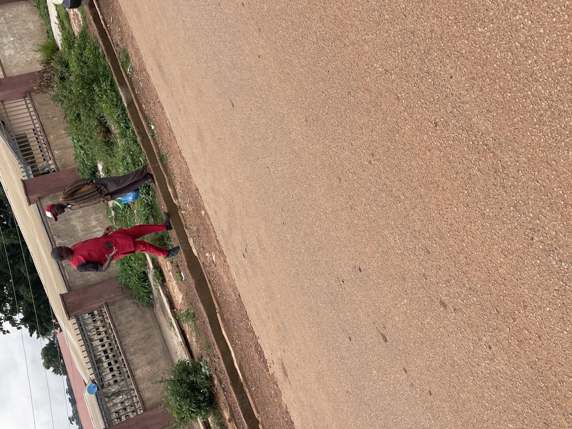In Gura Top, Nigeria, two individuals are the focus of this media. One person, wearing a vibrant red outfit with black accents on the sleeves and a red cap, stands on a paved surface. Their posture suggests they are in mid-conversation or explaining something, with one hand raised. Beside them, another person, dressed in a patterned top and dark trousers, stands in a more relaxed pose, holding a blue object.
The scene appears to be an outdoor setting, likely a street or pathway, with a building visible on the left side. The building has a weathered concrete facade and metal bars over windows or openings. Lush green vegetation lines the edge of the paved area, suggesting a natural environment contrasting with the urban elements. A drainage channel runs alongside the pavement. The ground is covered with a reddish-brown asphalt or concrete material, indicative of a warm climate. The lighting suggests it is daytime, possibly overcast as the sunlight is diffused. The overall mood is neutral, focusing on the individuals and their surroundings. No visible text is present in the image.
