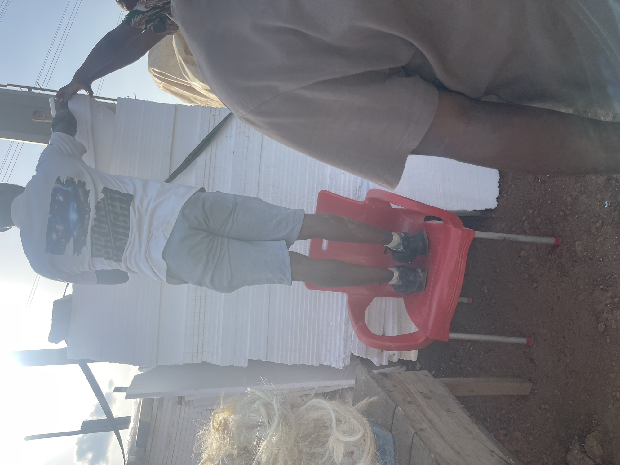 The image shows two people working with styrofoam sheets. One person stands on a red plastic chair, while the other is partially visible. The environment appears to be a construction or work site with dirt ground and wooden planks visible. The location is unknown, I have no evidence that it is Abuja, Nigeria.
