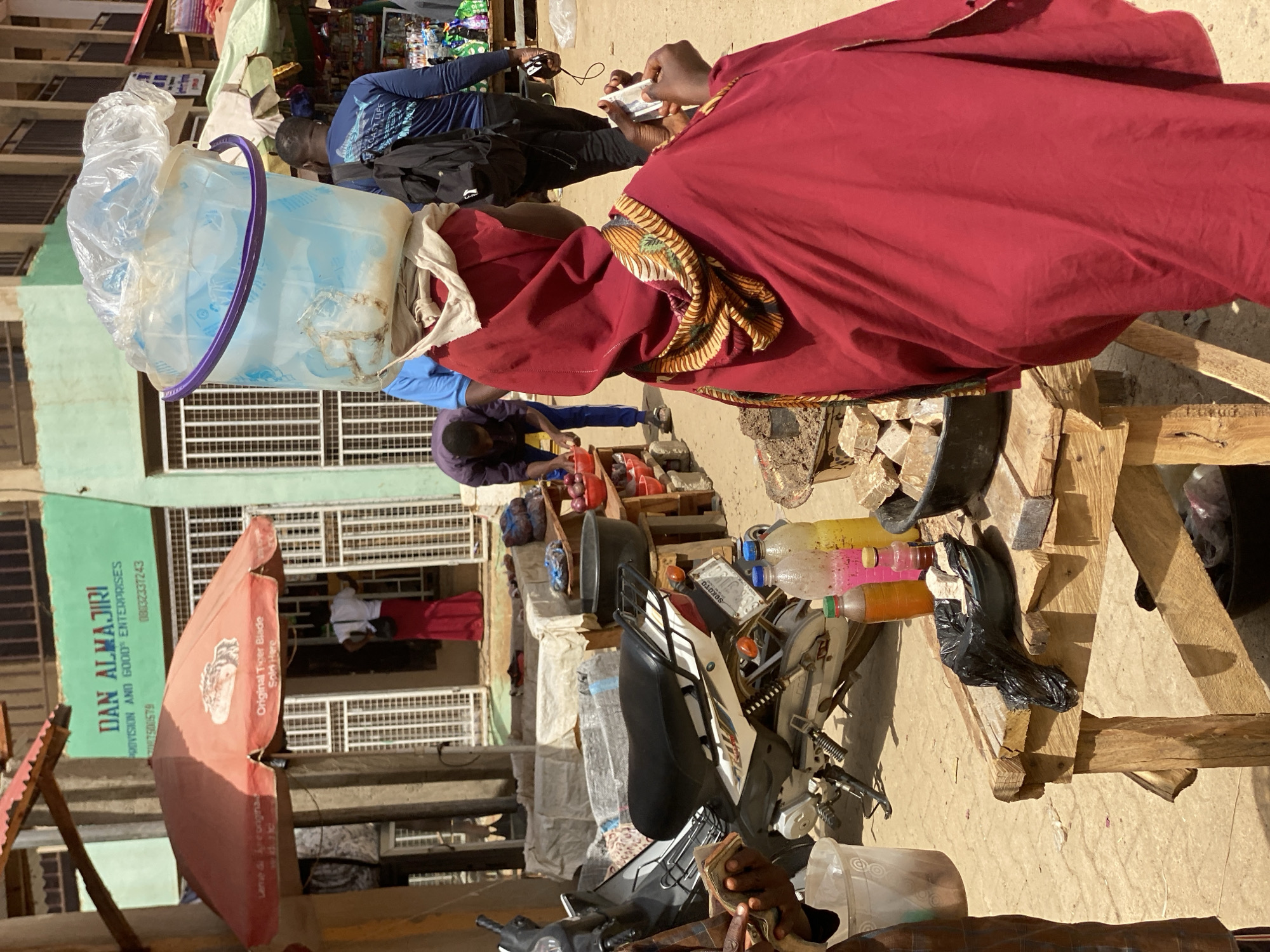 This sunlit image captures a bustling street market scene in Kafanchan B, Nigeria, during daytime. The scene is filled with people, market stalls, and various goods, indicating an active commercial environment.
In the midground, a person wearing a prominent red robe with a patterned trim on the sleeve is seen from behind, holding a small white object, likely a phone. To their left, another individual in a blue shirt holds a black object, possibly another phone. Further back, a person in a blue long-sleeved top is engaged near a market stall, and another individual in a purple top is bending over, seemingly inspecting items. In the background, a person in a red outfit stands near a building entrance.
Notable objects include a large, translucent light blue plastic bucket, partially covered by a clear plastic bag, which appears to be carried on someone's head. A black and white motorcycle is parked with various items on and around it, including several small plastic bottles containing colorful liquids (possibly beverages or oils) displayed on a wooden stand. A large red market umbrella, open for shade, stands prominently. Makeshift wooden structures and crates are used as market stalls or display areas.
The bright, direct sunlight suggests it is mid-day with clear weather. A light green building forms part of the backdrop, featuring a white grilled window. Visible text on the green building sign reads "DAN ALMAJIRI PROVISION AND GOOD ENTERPRISES" along with two phone numbers, "08032337243" and "7500579". The red umbrella clearly advertises "Original Tiger Blade Sold Here." The word "SOKOTO" is also visible on what appears to be a motorcycle's license plate. The overall activity suggests everyday commerce and social interaction within the market.