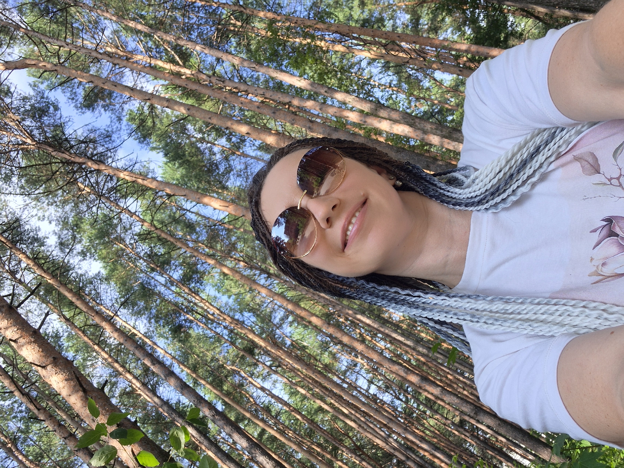 The image shows a woman with braided hair, wearing sunglasses and a white t-shirt, posing for a selfie in a forest. The background consists of tall pine trees with a bright blue sky visible through the canopy. The trees are angled, suggesting the photo was taken from a low angle looking upwards. The woman's braids are a mix of white and grey tones. The lighting indicates it is daytime, likely a sunny day. The location is implied to be a forest in Chaikovsky, Russia, based on the prompt's context.