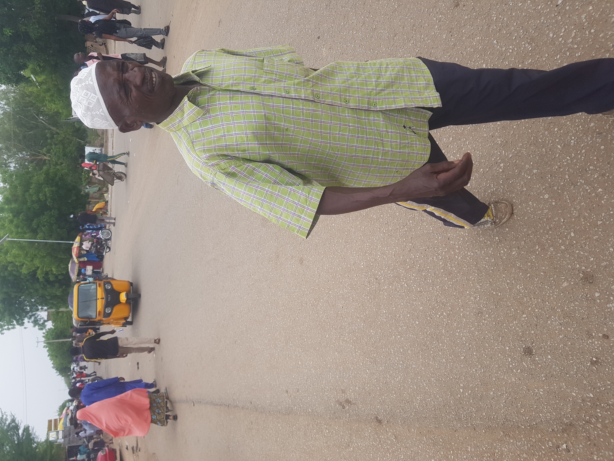 An elderly man wearing a white kufi cap and a plaid, short-sleeved green shirt stands on a paved road in Dabawa, Nigeria. He wears dark trousers with a yellow stripe near the hem and beige shoes. Other people walk in the background. A yellow tricycle taxi is parked on the side of the road. Trees and buildings are visible in the background. The road appears to be a main thoroughfare.