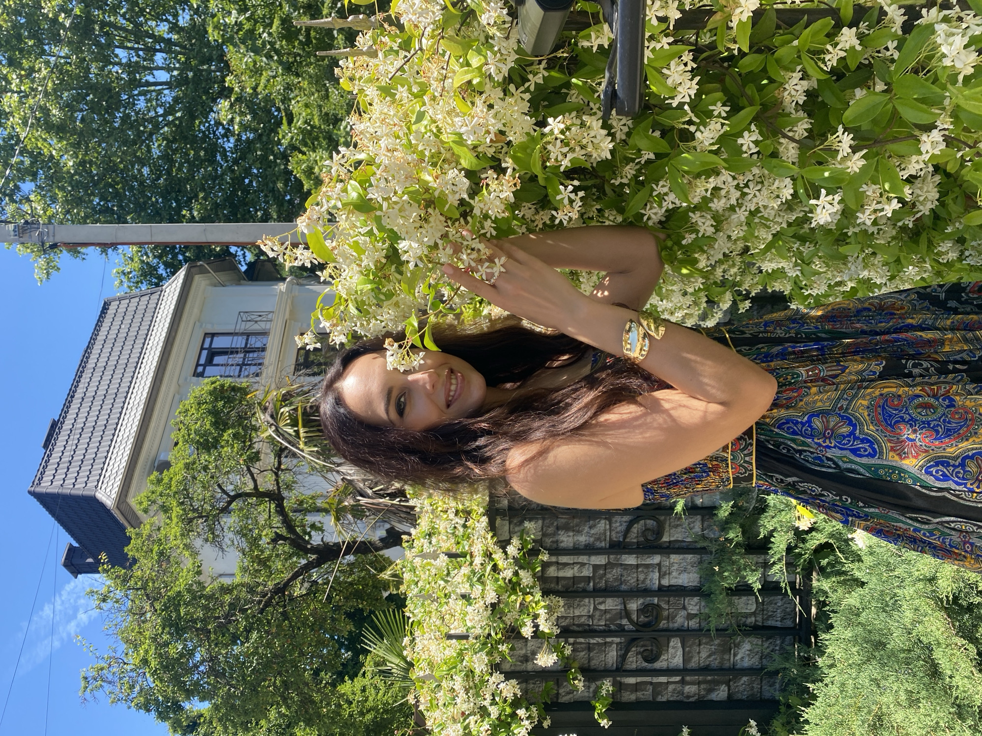 A medium-shot, eye-level, outdoor, daytime, vertical photograph shows a woman with long, dark hair standing in front of a wall and a blooming vine. The woman is smiling and looking at the camera. She is wearing a colorful, patterned dress and a gold bracelet. The vine is covered in small white flowers. In the background, there is a white house with a dark roof and a black fence. The location is the city of Sochi, Russia.