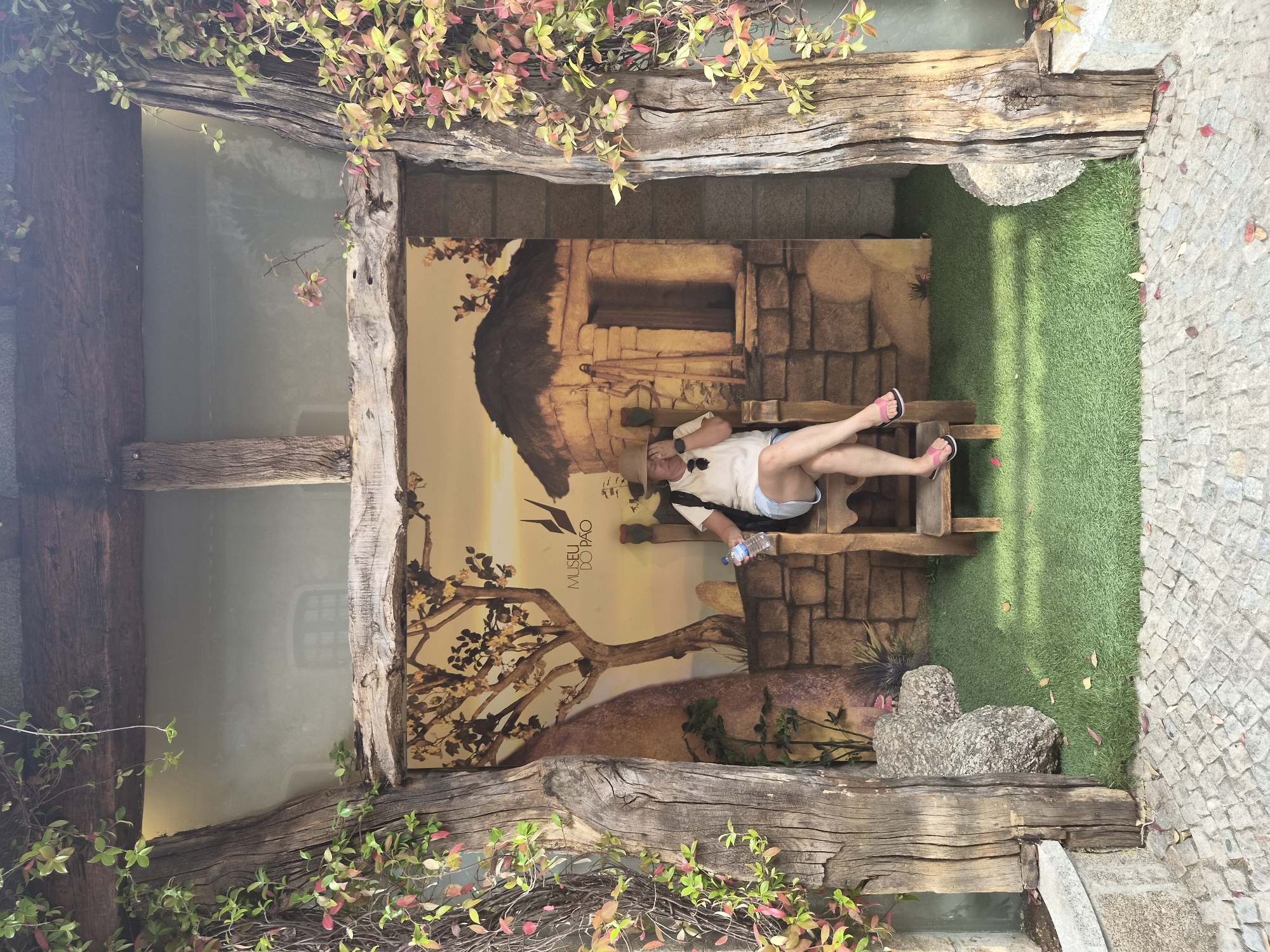 The image depicts an outdoor scene in Seia, Portugal, featuring a woman seated on a wooden chair. She is wearing a straw hat, a white t-shirt, and denim shorts, and is holding a bottle of water. The backdrop behind her is a mural that resembles a rustic village scene with a stone building, a tree, and a sunset. The scene is framed by rustic wooden pillars entwined with ivy, suggesting an entrance or a decorative feature. The ground is covered with artificial grass in front of the mural and cobblestones leading towards the viewer. The overall atmosphere appears to be a tourist attraction or a themed exhibit.