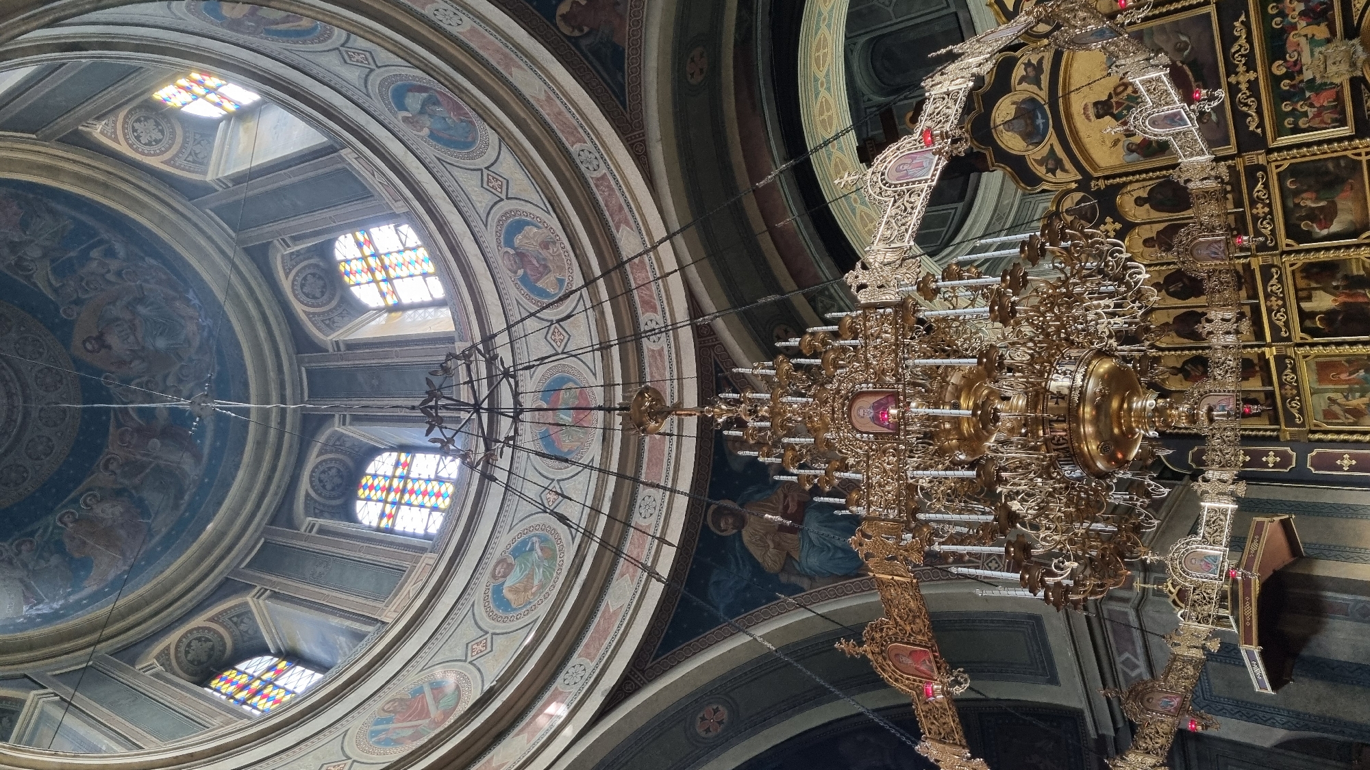 This is an interior view of a domed church, likely a cathedral, in Chernivtsi, Ukraine. The ornate golden chandelier suspended from the ceiling is the central focus. The dome is decorated with religious paintings, possibly frescoes, depicting figures in halos. Several stained-glass windows, featuring multicolored panes, are visible around the base of the dome, letting in light. The walls below the dome are also adorned with religious imagery, including a large painting of a seated figure holding a book. Below and to the right of the chandelier, a large iconostasis, a wall of religious icons, is partially visible, decorated with gilded frames and paintings. The overall impression is one of grandeur and religious devotion.