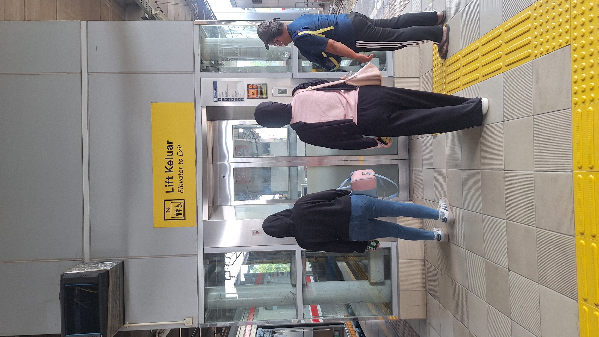 People wait at an elevator in a public transport station in Cikarang, Indonesia. A person in a blue shirt and dark pants stands at the end of a tactile paving strip, looking into the elevator. Two people stand in front of the elevator doors. One person wears a pink and black outfit with a head covering, while the other person wears a black hoodie and blue jeans. A yellow sign on the left of the elevator doors reads "Lift Keluar Elevator to Exit" with symbols indicating accessibility for wheelchairs and people with strollers.