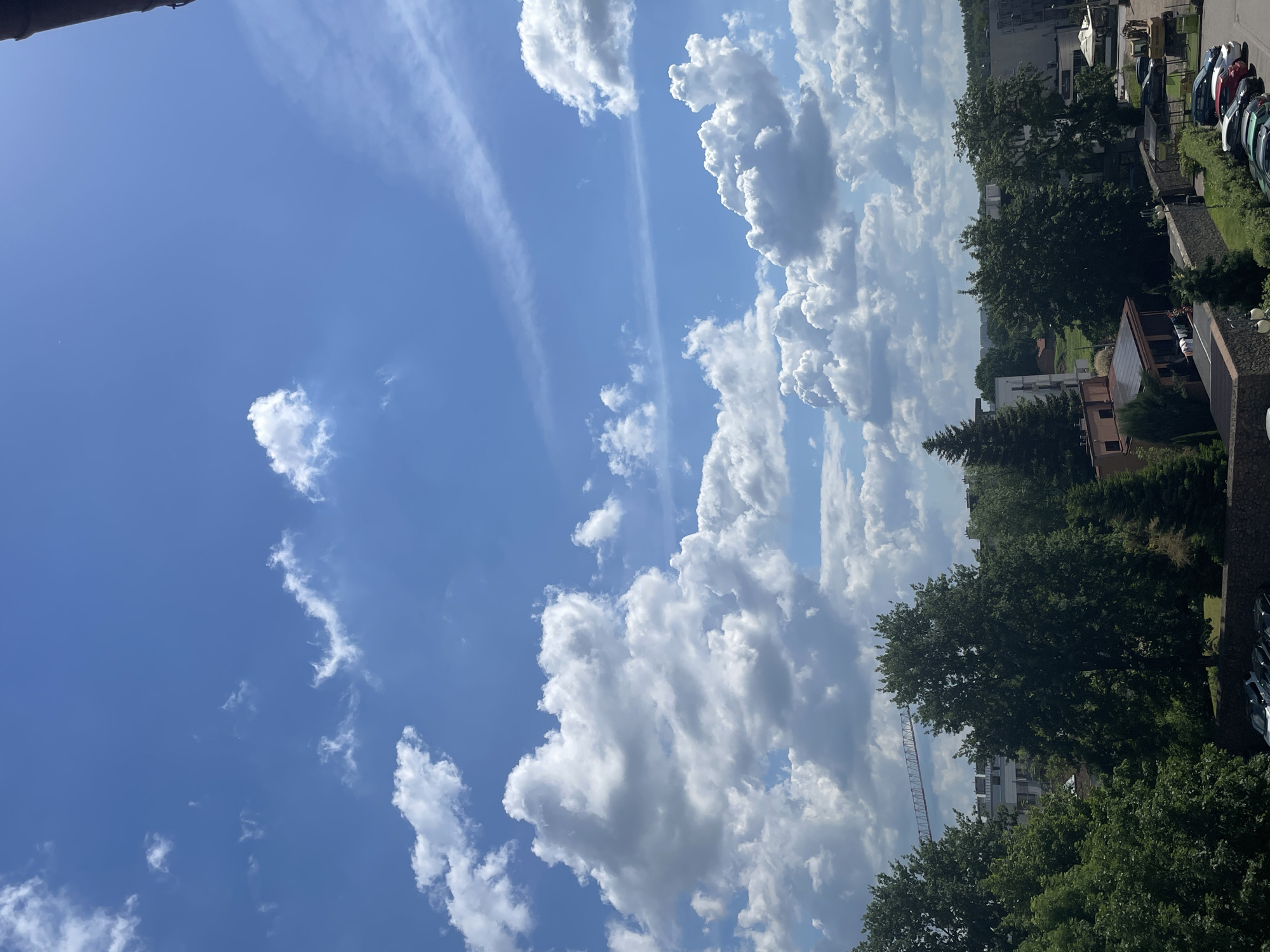 The image shows a cityscape under a partly cloudy blue sky. The foreground features a row of trees and some buildings. In the middle ground, there are more trees, buildings, and a construction crane in the distance. The sky is dominated by fluffy white clouds against a bright blue backdrop. The lower part of the image shows a parking lot with several cars.