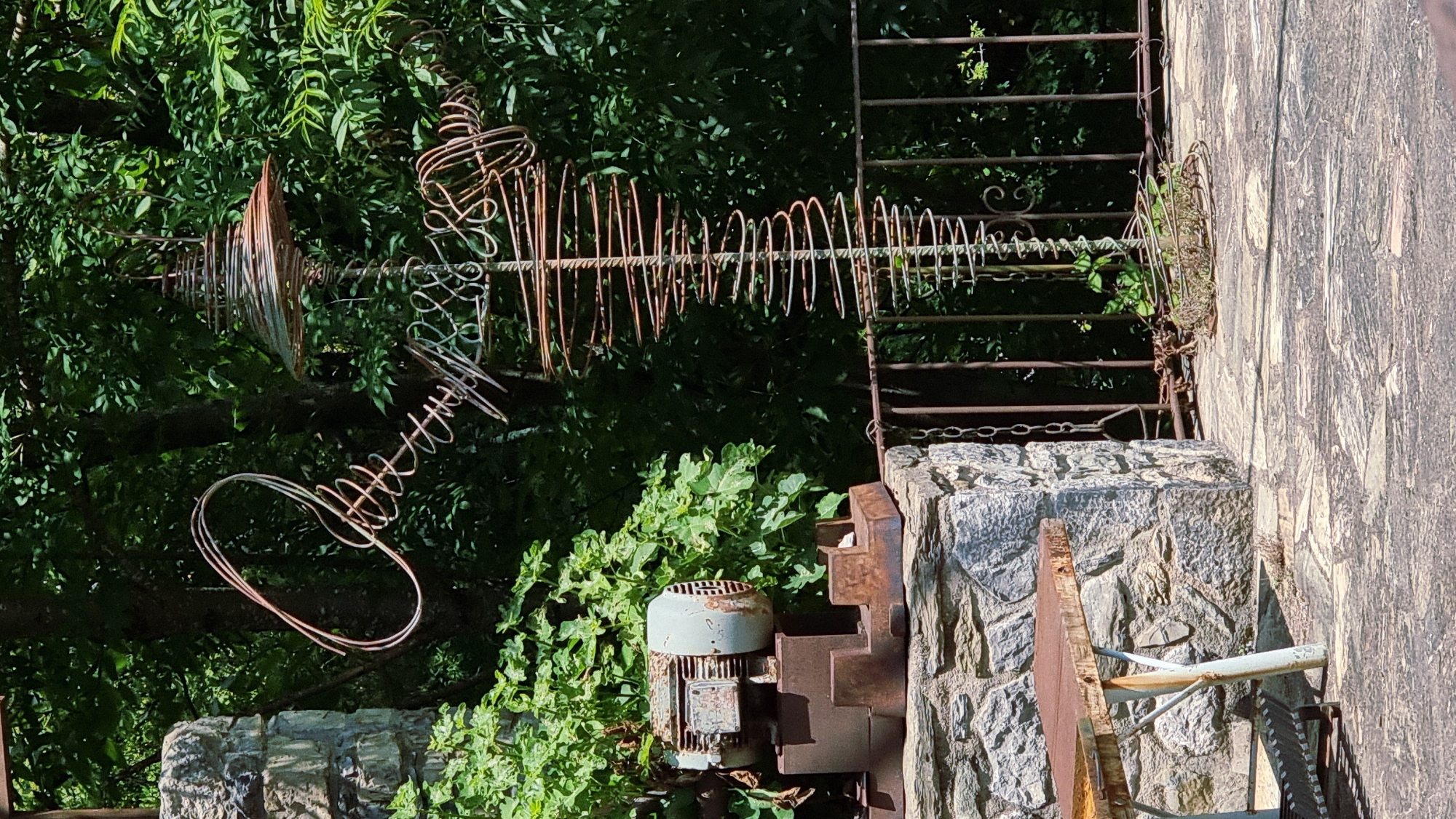 The image shows an outdoor scene in Camprodon, Spain, featuring a wire sculpture in the shape of a human figure. The sculpture is made of rusty wire and is positioned in front of lush green foliage. To the left of the sculpture, a stone structure is visible, topped with a rusty industrial motor. A metal bench with a rusty tabletop stands in front of the stone structure. A rusty metal railing with chains runs along the edge of the paved area where the sculpture is located. The ground is paved with flagstones. The overall scene appears to be an artistic installation in a garden or outdoor space.