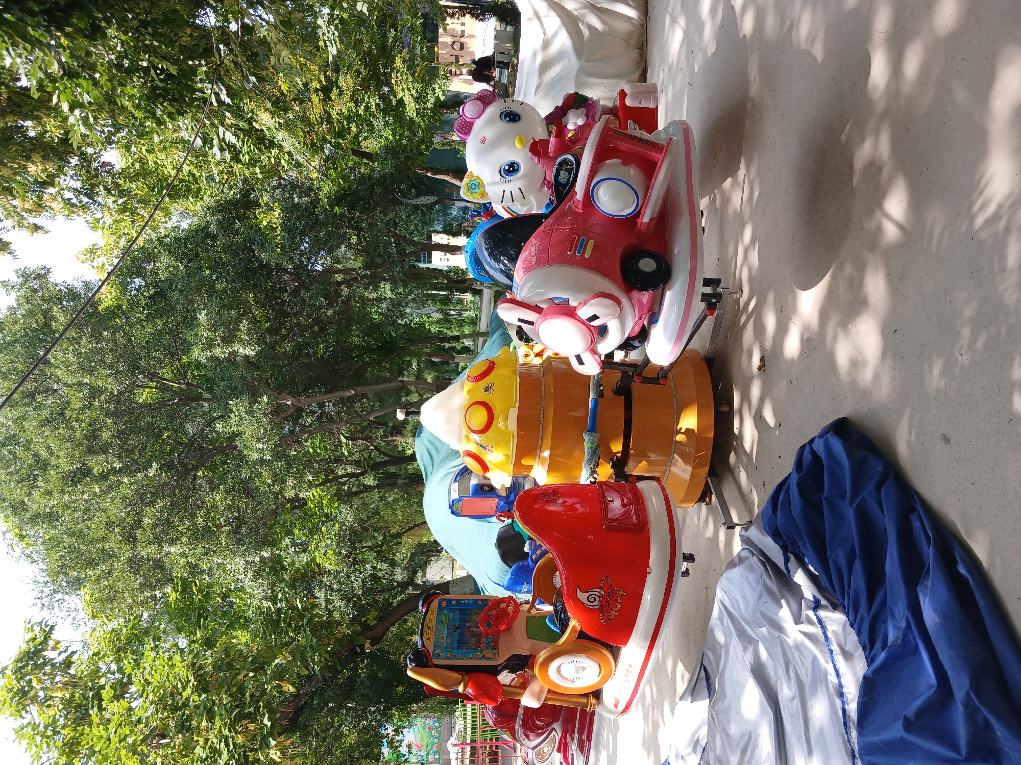 The image shows a line of brightly colored children's amusement rides, possibly in a park or outdoor play area. The rides are designed as various vehicles and characters, including a pink airplane with Hello Kitty as the pilot, a yellow barrel-shaped ride, and a red motorcycle with a cartoon character. The rides are lined up on a paved surface, with trees and greenery visible in the background, suggesting an outdoor setting. The sunlight filtering through the leaves creates dappled shadows on the ground.
The image appears to have been taken during the daytime, with bright sunlight and clear skies, as indicated by the strong shadows. The overall mood is cheerful and playful, due to the vibrant colors of the rides and the presence of children's entertainment. There is no specific activity or event depicted, other than the rides themselves being stationary.
The location cues, such as the type of trees and the general ambiance, are consistent with a park or amusement area in Tashkent, Uzbekistan. Some of the rides have text and numbers, but they are not clearly legible in the image. For example, the pink airplane has "000" visible on its side. The ride in the foreground has some indecipherable text and graphics.