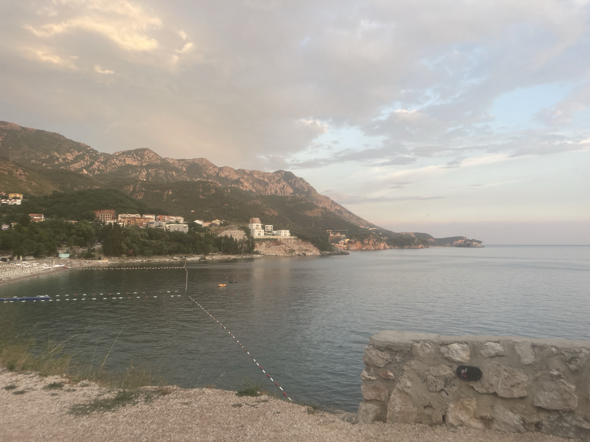 The image depicts a serene coastal scene at Kamenovo, Montenegro, likely during the late afternoon or early evening. The sky is partly cloudy with soft, diffused light, suggesting either an approaching sunset or a generally overcast day. The sea is calm and stretches out to the horizon, meeting a pale sky.
In the foreground, a low, rough stone wall lines the edge of a gravelly embankment, with some sparse grass growing near it. Beyond the wall, a beach is visible, lined with numerous white lounge chairs and umbrellas, indicating it's a tourist or resort area. The water near the shore is shallow and marked with buoys and ropes, delineating swimming areas. There are a few small objects in the water, possibly buoys or inflatable toys. A couple of small boats or watercraft are also visible.
Further back, nestled against the foothills of a mountain range, are several buildings. These appear to be hotels or residential complexes, with white and earth-toned facades. The mountains themselves are rugged and covered in green vegetation on their lower slopes, becoming more rocky and bare towards their peaks. The overall mood is peaceful and tranquil, with a sense of natural beauty and leisure. No visible text is present in the image.