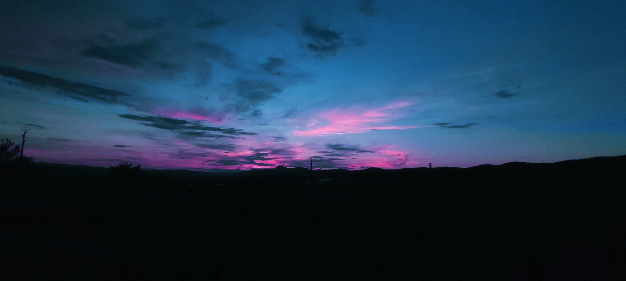 A wide-angle, low-light landscape photograph captures a dramatic twilight sky over Momin Prohod, Bulgaria. The sky is dominated by vibrant hues of deep blue, transitioning to streaks of electric pink and purple, indicating the lingering light of sunset or the approaching dawn. Wispy, dark clouds are scattered across the upper portion of the sky, contrasting with the brighter colors. The horizon is defined by a silhouetted mountain range, appearing as a dark, undulating line against the colorful sky. In the foreground, the land is completely dark, obscuring any details of the terrain or any structures or vegetation that might be present. A single, dark utility pole is visible to the left of the frame, and another smaller pole or structure is barely discernible on the right. The overall impression is one of natural beauty and the ephemeral quality of twilight.