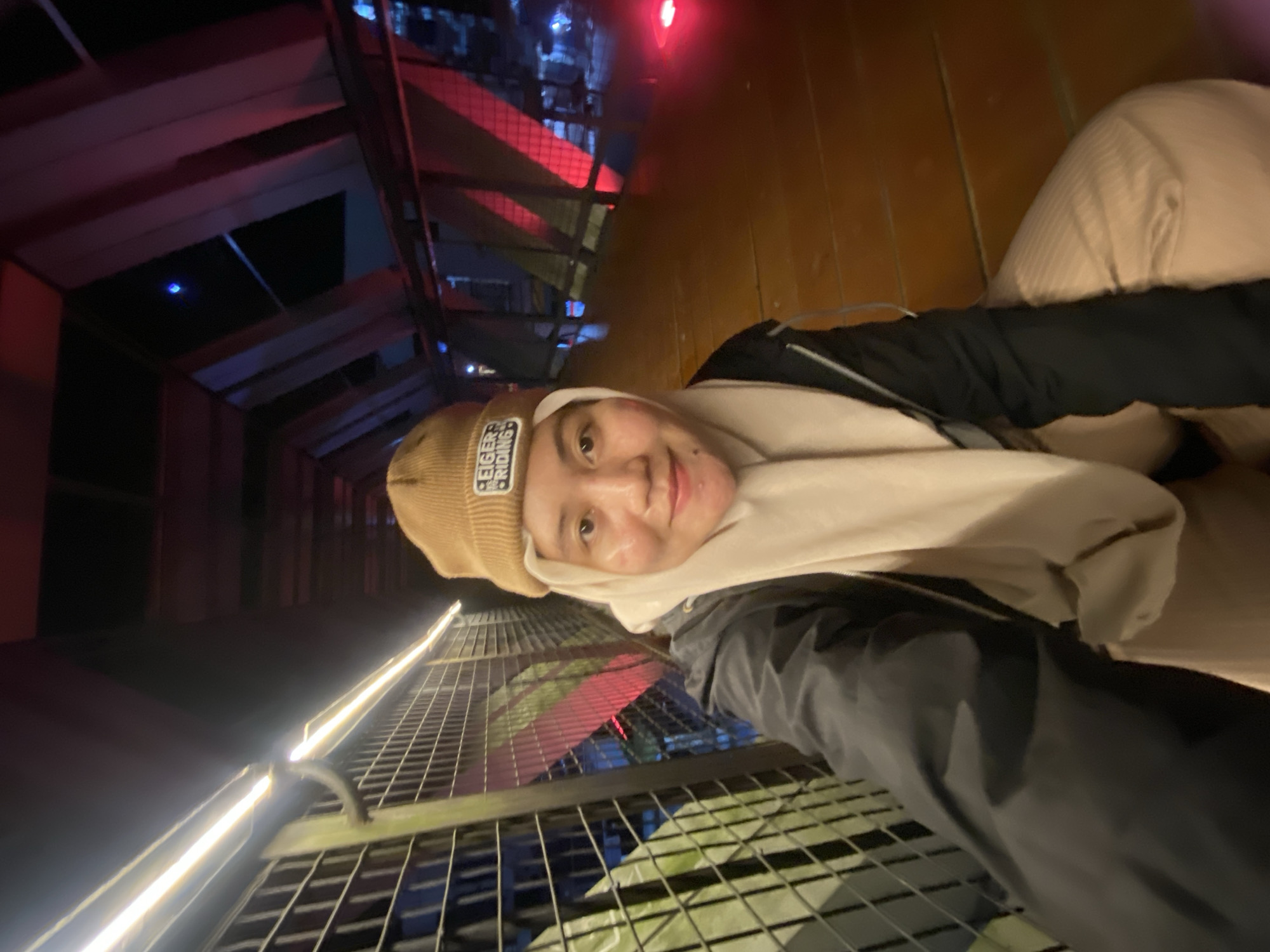 This is a selfie taken by a person wearing a tan beanie and a light-colored scarf or hoodie. The person has a gentle smile. They are wearing a black jacket over a light-colored garment. The background shows an indoor or covered walkway with a metal grate floor and railing. There are illuminated lines along the walkway and some reddish-pink lighting in the distance, suggesting an entertainment venue or a pathway with decorative lighting. The time of day appears to be evening or night due to the artificial lighting. The texture of the wooden wall on the right side of the image is visible. The text "EIGER RIDING" is visible on the beanie. The location context is Banjarbaru, Indonesia.
