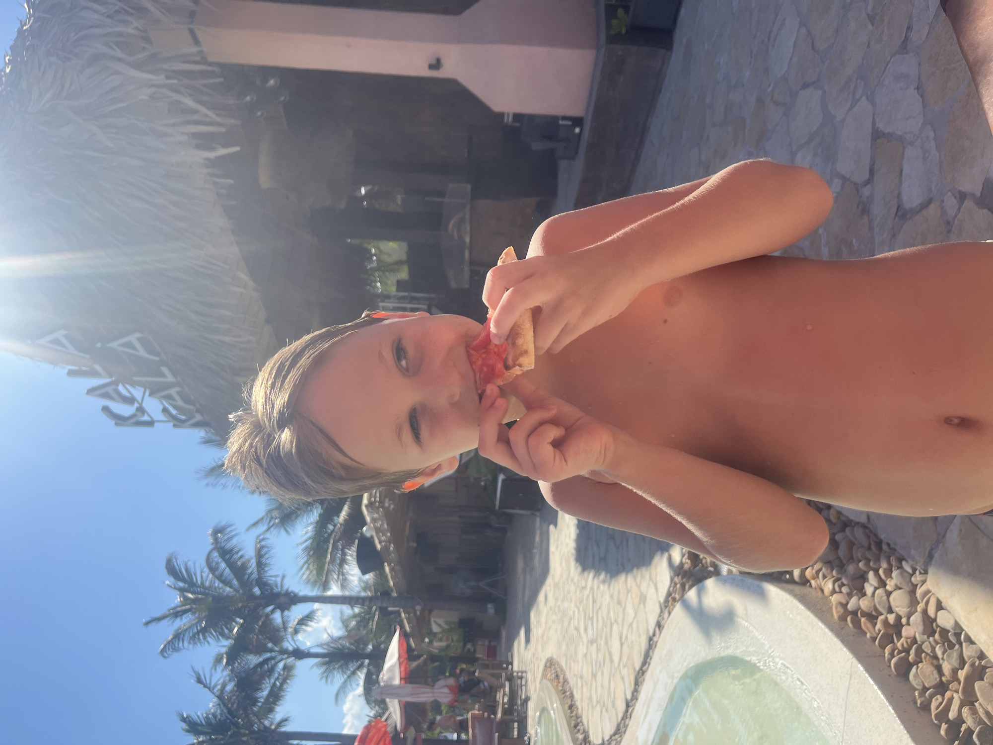 The image depicts a young child, likely a boy, heartily eating a slice of pizza in a vibrant, outdoor setting in Đà Nẵng, Vietnam. The child, who is shirtless, holds the pizza close to his face, taking a bite and looking towards the camera with a contented expression.
The scene is set in what appears to be a tropical resort or pool area. In the background, tall palm trees with green fronds stand out against a clear, brilliant blue sky, indicating bright and sunny weather. A large thatched-roof structure, possibly part of a bar or shaded seating area, is prominent on the left, contributing to the tropical aesthetic. The ground is paved with irregular stone tiles, and next to the child, a small, inviting body of water, resembling a hot tub or a small pool, is visible, surrounded by a border of smooth pebbles.
The bright sunlight suggests it is midday or early afternoon, casting distinct shadows and creating a slight lens flare on the left. Other elements in the background include more resort architecture and faintly visible outdoor furniture, hinting at a leisurely atmosphere. The child's happy demeanor while enjoying his snack perfectly encapsulates a relaxed vacation moment. There is no visible text in the image.