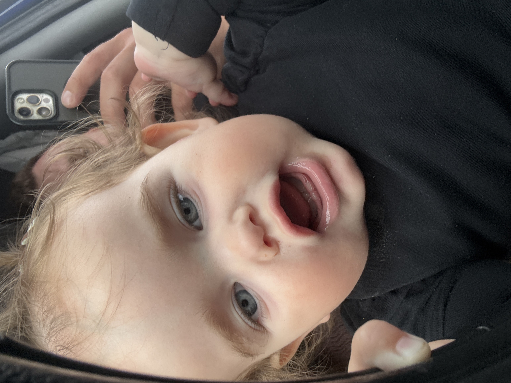 The image is a close-up, selfie-style photograph of a baby's face. The baby has fair hair, blue eyes, and rosy cheeks. Their mouth is open, as if speaking or laughing. The baby is wearing a black long-sleeved shirt. In the background, a hand is visible holding a smartphone, which is reflecting the scene. Another person's head is also partially visible behind the baby. The lighting suggests an indoor setting, possibly a car due to the reflections. There is no indication of the location being in Koblenz, Germany.