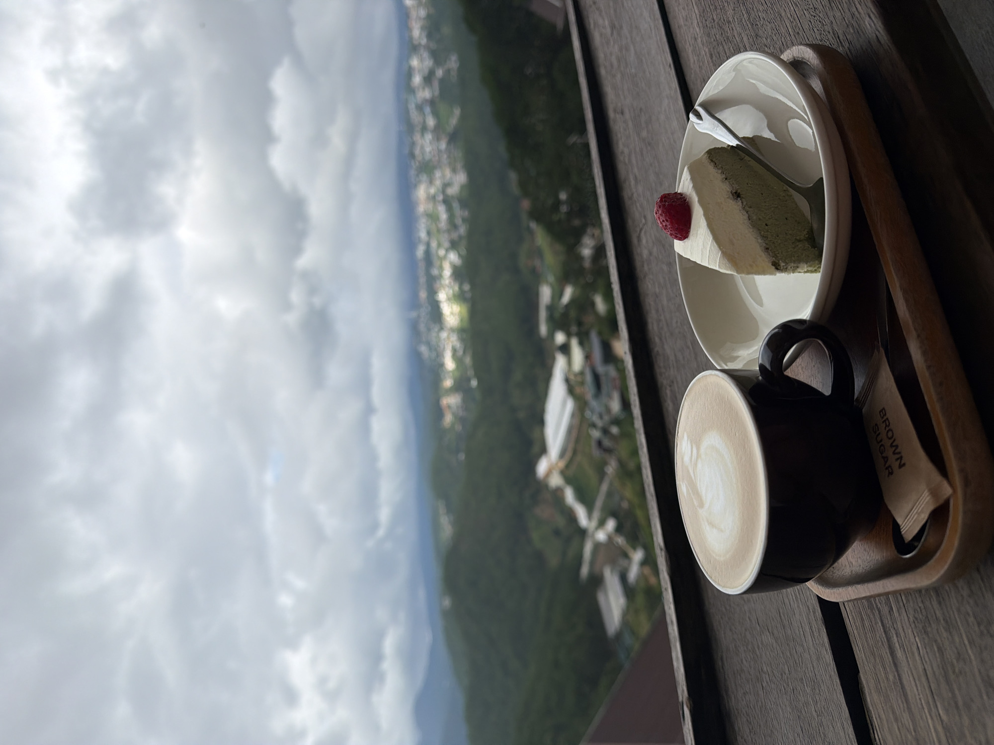A slice of cake with a strawberry on top sits on a white plate next to a cup of latte art coffee, both placed on a wooden tray. The tray is on a dark wooden table. In the background, a hazy cityscape with rolling green hills under a cloudy sky is visible. The scene is likely from a cafe or restaurant in Xuân Thành, Vietnam, offering a scenic view.