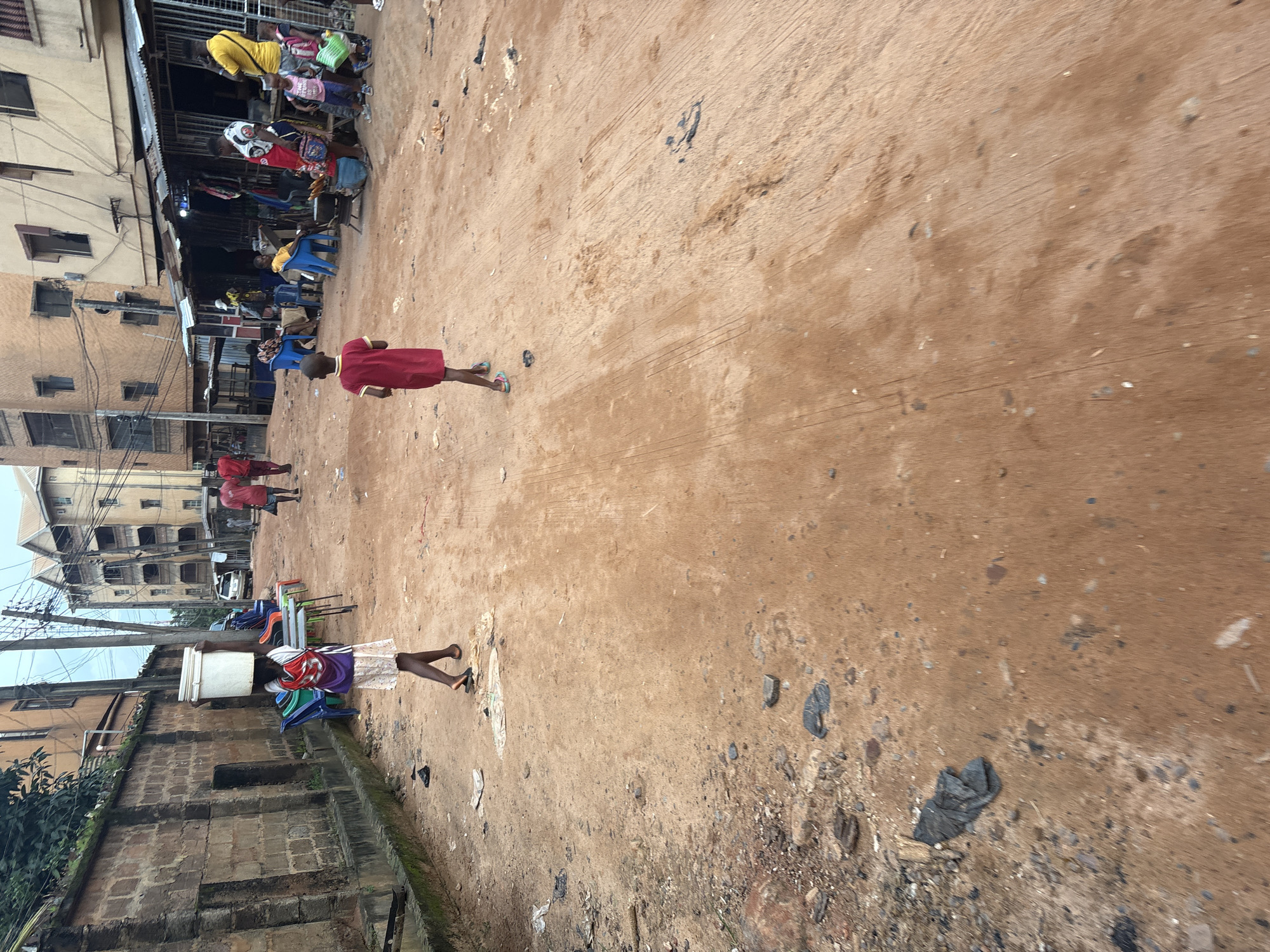 In Nkpor, Nigeria, a young person walks down a dirt road, carrying a large white bucket on their head. They are wearing a white shirt with a red and purple design, a purple vest, and a light-colored skirt. The road is dusty and uneven, with scattered debris. Buildings line the street, some appearing to be shops with corrugated metal roofs. Several other people are visible in the background, some sitting on plastic chairs outside the shops, while others are walking or standing. A few vehicles are parked along the roadside. The scene depicts a typical day in a bustling urban environment in Nigeria.