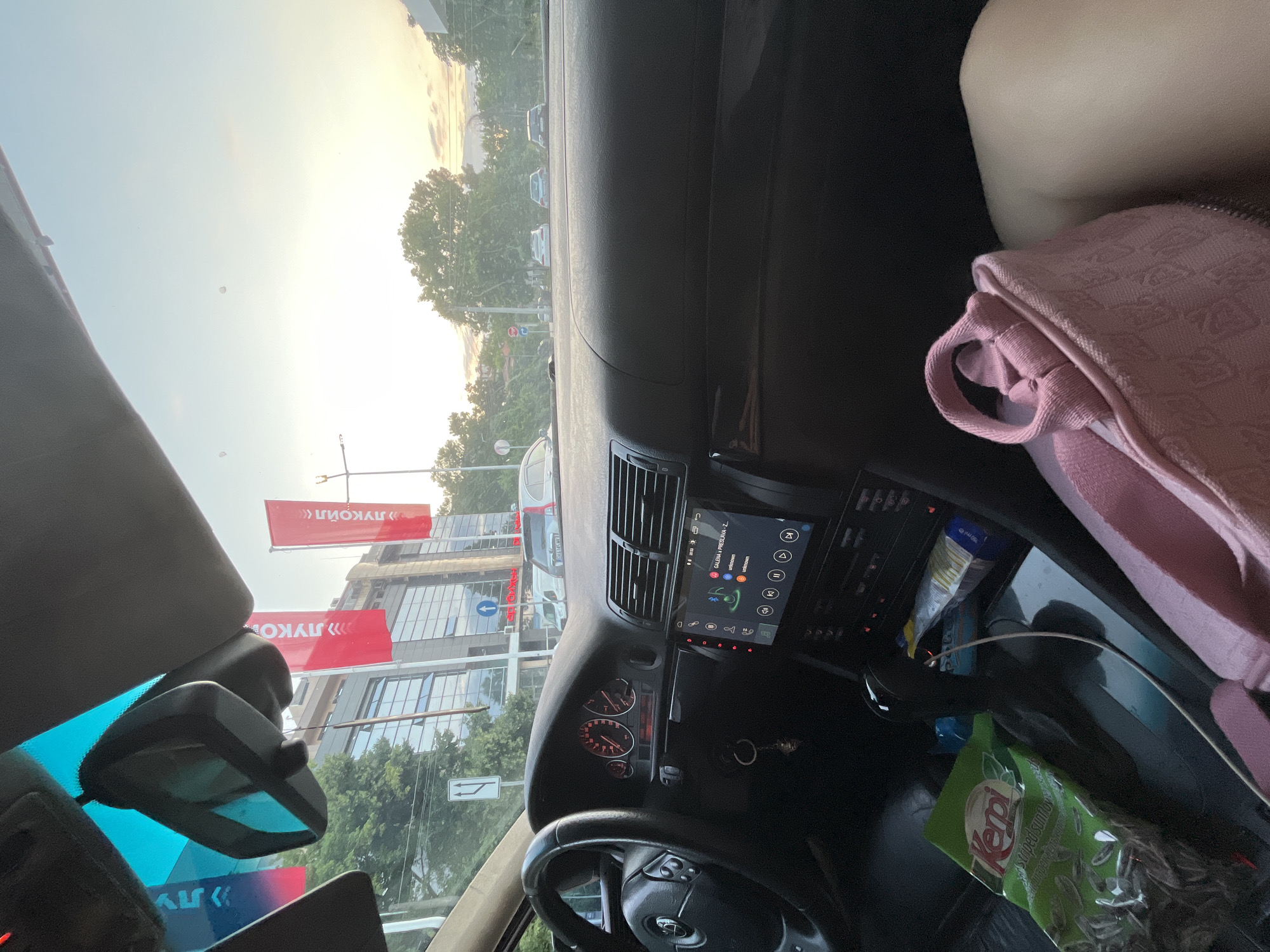 The image is taken from inside a car, looking out through the windshield and side windows. The car's dashboard is visible, including the steering wheel, instrument cluster, and a modern infotainment screen. A bag of "Kepi striped sunflower seeds" and a pink fabric bag are on the center console. Red banners with Cyrillic text, likely advertising, are visible outside, along with a building with many windows. Several cars are on the road, and trees and a street light are also present. The sky suggests it is late afternoon or early evening, with a sunset glow.