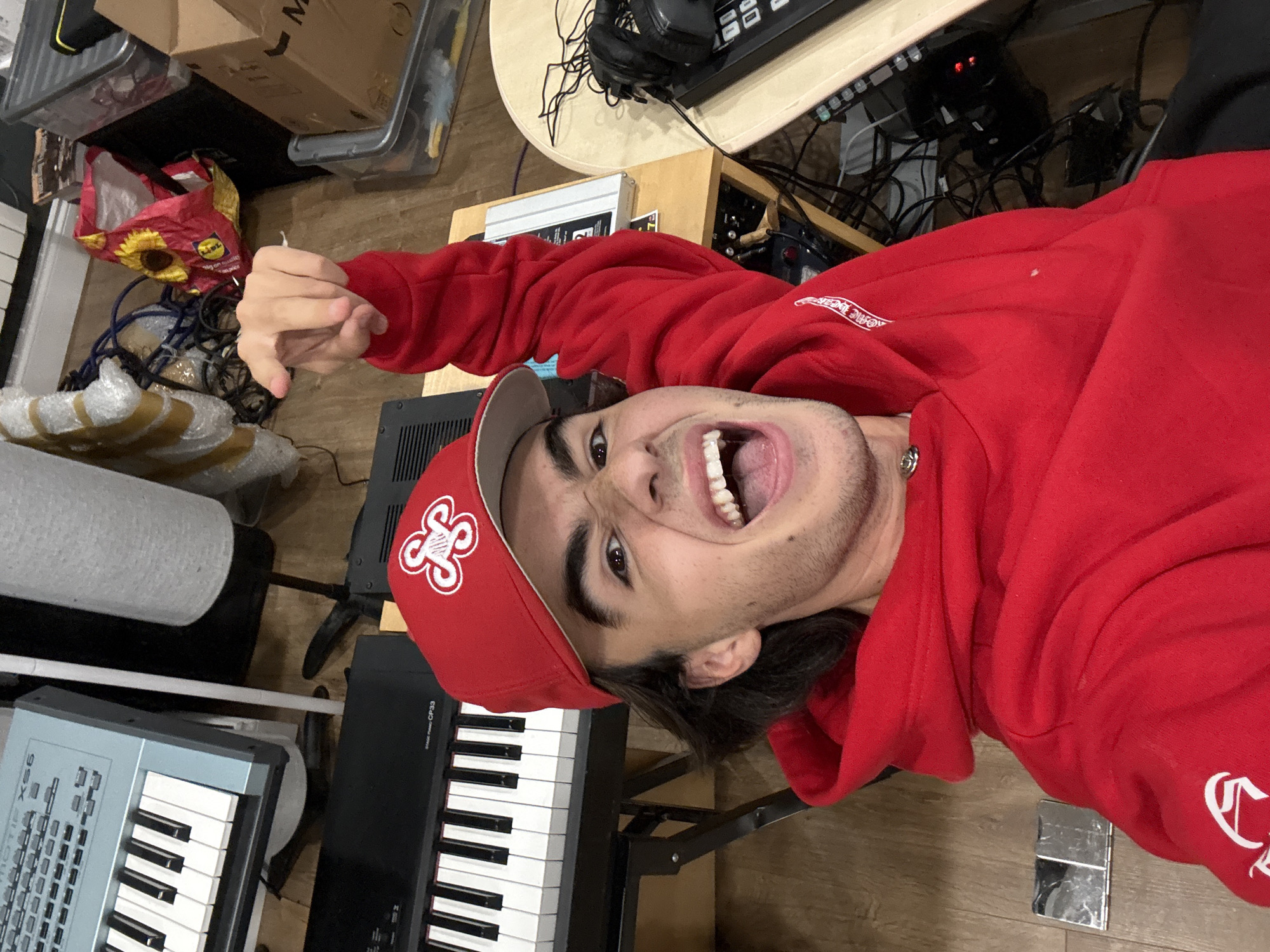 A young person, wearing a red hooded sweatshirt and a red baseball cap, is pictured in a room with musical equipment. The person is smiling with their mouth open and pointing their right index finger towards the viewer. In the background, there are several keyboards, including a Yamaha MOTIF XS6 and a Yamaha CP33 Stage Piano, along with various cables, boxes, and a lamp. The lighting suggests an indoor setting, possibly a home studio. The overall impression is one of a vibrant and creative space.