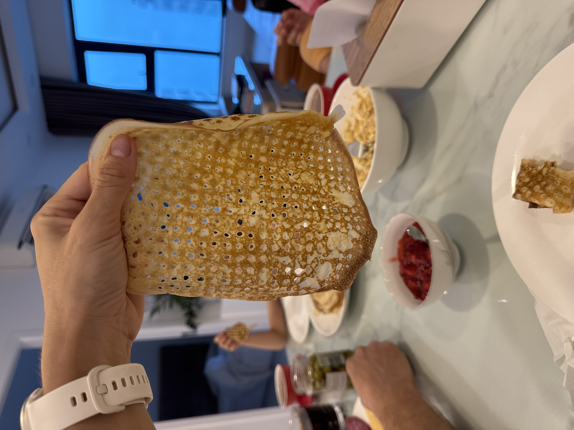 A hand holds a thin, porous pancake or crepe. Bowls containing toppings such as fruit or syrup and a small dish of rolled crepe pieces are on a light colored tabletop. The image shows other people seated at the table. Utensils and condiments are scattered on the table. A window showing a blue sky and trees in the background suggests an interior setting. The location is Bac Nha Trang Ward, Vietnam, based on the GPS information.