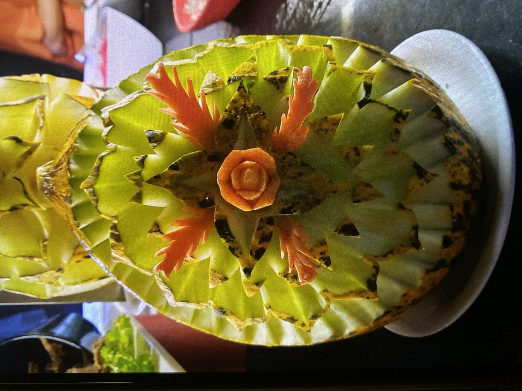 An elaborately carved fruit, possibly a large melon, is the central subject of the image, presented on a white plate. The fruit's rind is intricately cut into numerous overlapping, serrated green segments, which feature distinctive dark, mottled edges, creating a textured and somewhat weathered appearance.
Adorning the surface of the carved fruit are several smaller, vibrant orange decorative elements. These include a central, delicate rose-shaped carving and multiple elongated, spiky, and flame-like carvings, which appear to be crafted from a different fruit or vegetable, such as a carrot or papaya.
The background is softly blurred, revealing hints of an indoor setting, likely a restaurant, hotel buffet, or catering event. Partial views of other dishes, including green leafy vegetables, are discernible, along with reflections from ambient or artificial lighting. The overall scene depicts a highly decorative food presentation, showcasing culinary artistry, possibly for a special occasion or meal service in Bankya, Bulgaria. No identifiable people or visible text are present in the image.