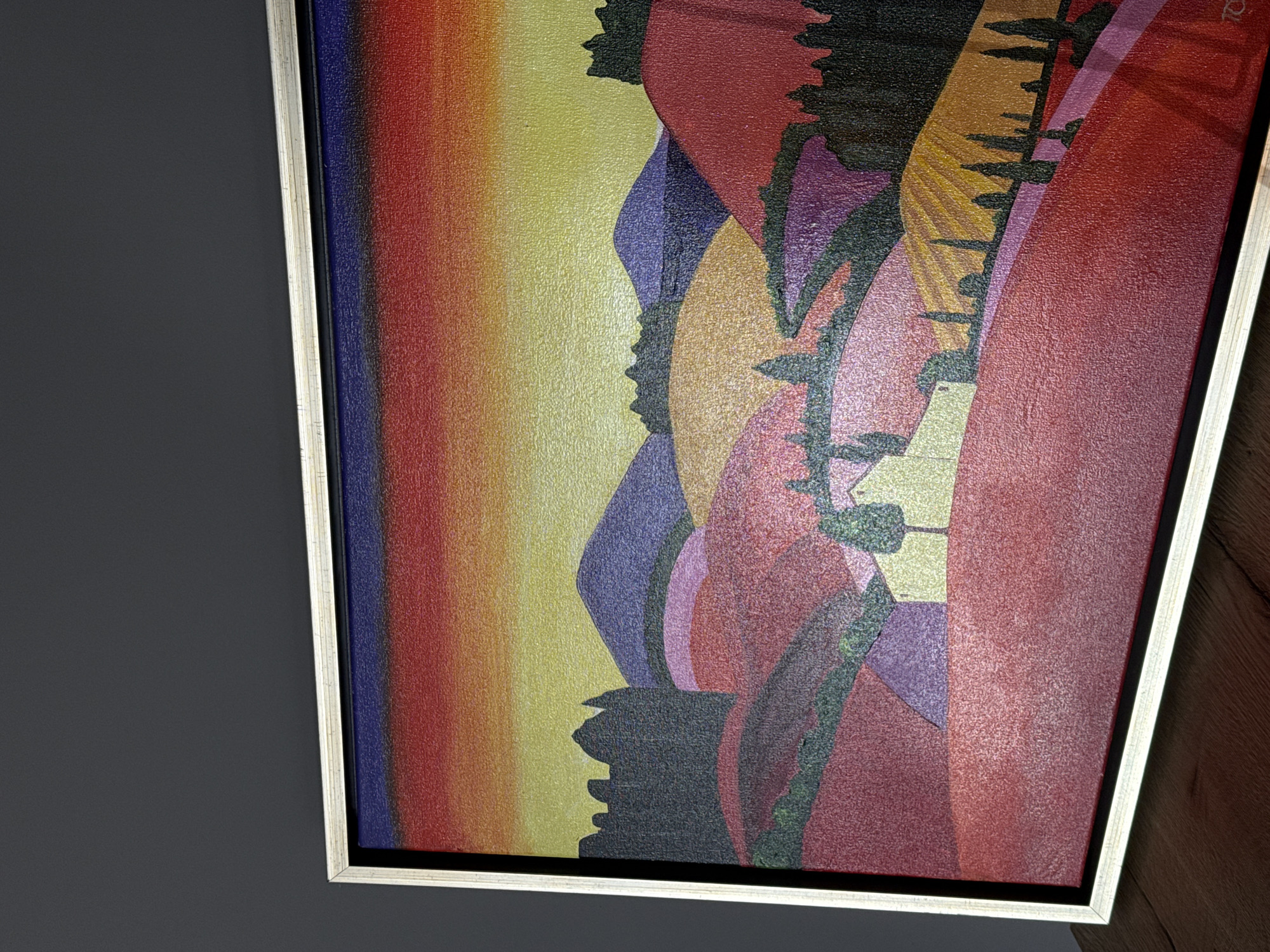 The image shows a framed painting of a stylized Tuscan landscape. The painting features rolling hills rendered in vibrant colors of red, purple, orange, and yellow, with a gradient sunset sky above. There are silhouetted trees, a building with a light yellow roof, and what appears to be a castle or fortress on the left side of the frame. The painting is displayed against a dark gray background and rests on a wooden surface. There is no indication that this image is related to the city of Trud, Bulgaria.