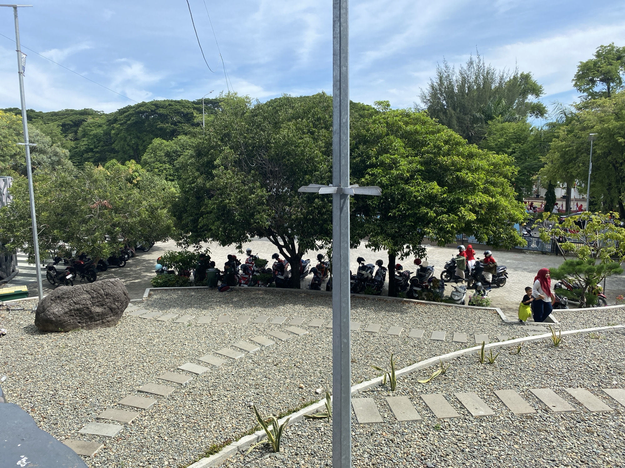 This is an outdoor scene in Banda Aceh, Indonesia, characterized by a parking area and a landscaped garden. In the foreground, a pathway made of stepping stones traverses a gravel-covered ground. Several small plants are visible along the edges of the pathway. In the mid-ground, a large, rough-textured boulder sits on the gravel. Adjacent to the boulder is a parking area populated by numerous motorcycles, with some people standing or sitting near them. The people include a woman in a red headscarf and a child in a green garment, walking together, and other individuals in the background who appear to be interacting with the parked motorcycles. The background is dominated by lush green trees, forming a dense canopy. A tall, metallic pole with mounting brackets is positioned prominently in the center of the image. The sky above is blue with scattered white clouds.
