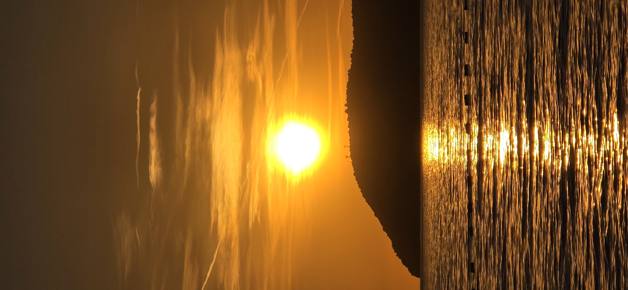The image depicts a sunset scene over the Adriatic Sea in Bar, Montenegro. The sun is a bright orb positioned centrally, with its light reflecting on the water's surface, creating vertical streaks of bright golden light. To the left of the sun, there is a diffuse orange sky with faint cloud-like patterns. A dark silhouette of a mountainous landmass separates the sky from the sea. The overall atmosphere is warm, dominated by shades of orange and yellow. There are some dark poles in the water.