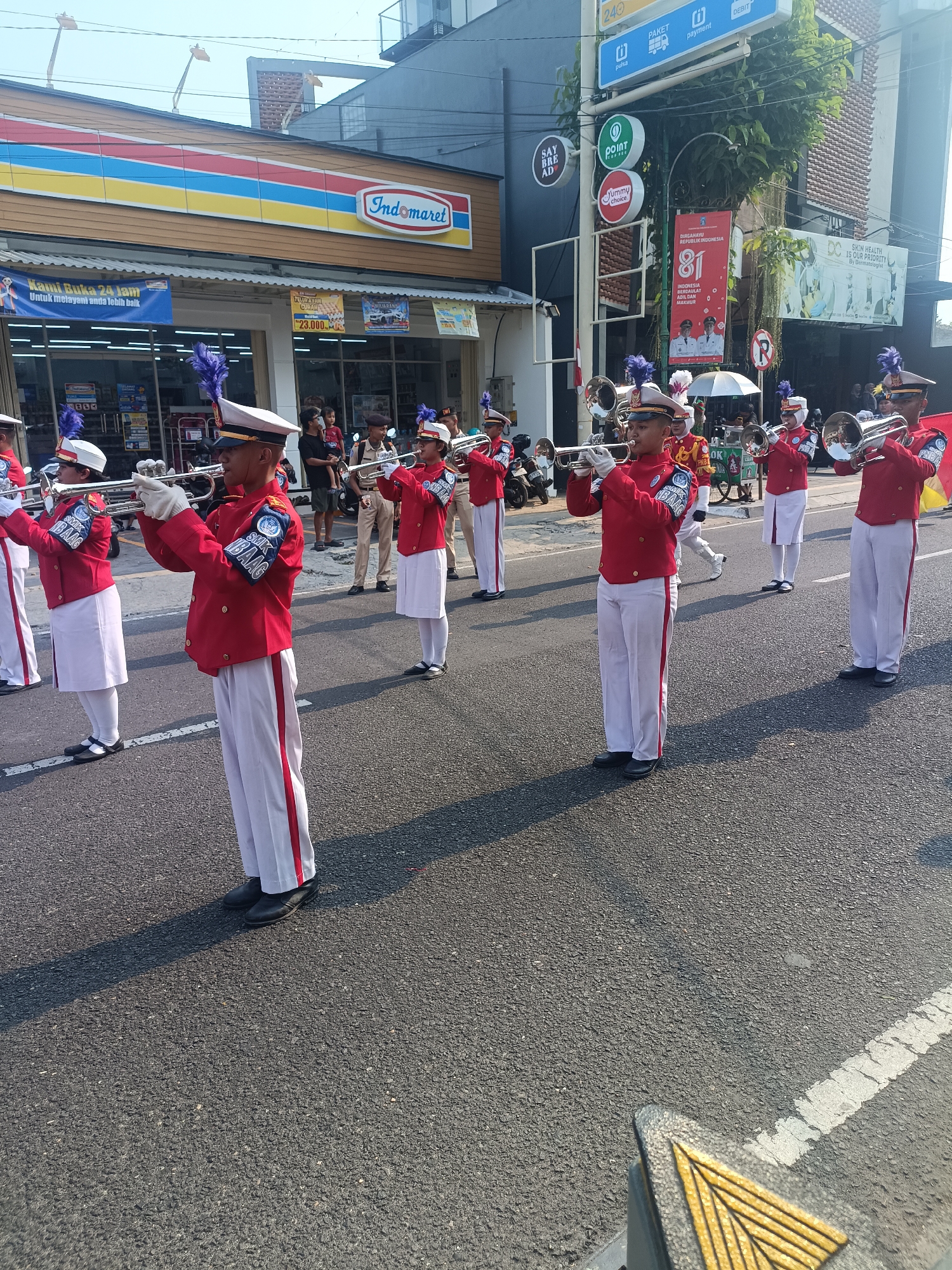 This is a medium shot, eye-level photograph captured on a street in Sleman, Indonesia, during the daytime. A marching band, composed of individuals in red jackets with white pants and hats adorned with blue feathers, is performing. They are holding and playing brass instruments, specifically trumpets. The band members are in formation, marching on an asphalt road. In the background, commercial buildings with signage are visible, including an Indomaret store and other shops. A traffic light and various advertisements are also present. The overall scene suggests a public event or parade.