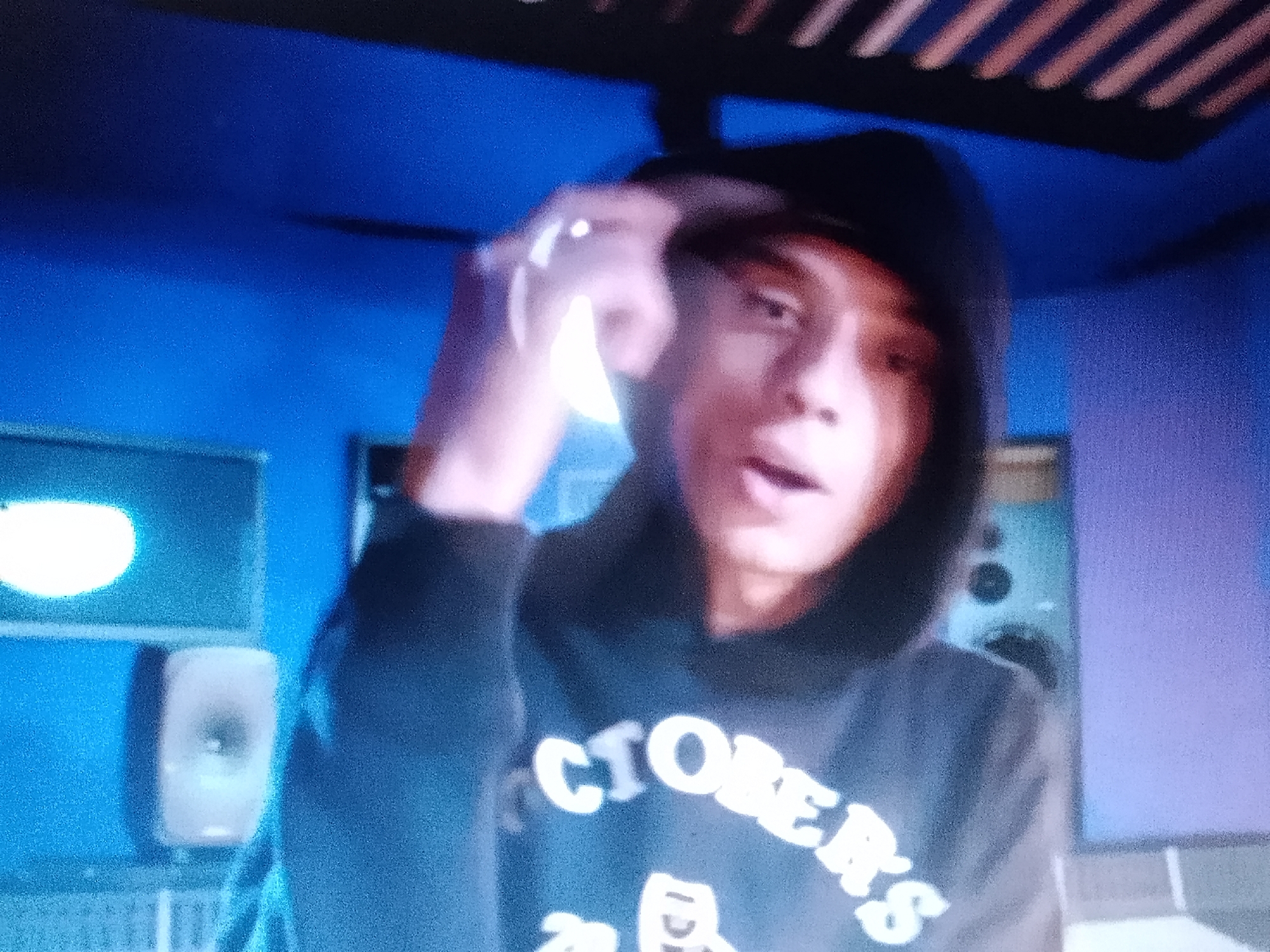 A medium shot captures a young individual wearing a dark hoodie with "OCTOBER'S" printed in a light color on the front. The person is holding their hand near their head, possibly adjusting something. The background is a blue-toned room with soundproofing panels on the ceiling and audio monitors visible on the left side of the frame. The lighting suggests an artificial source, creating a concentrated bright spot on the left and casting shadows in other areas. The image suggests an indoor studio setting. There is no information to indicate that the image was taken in Adankolo, Nigeria.