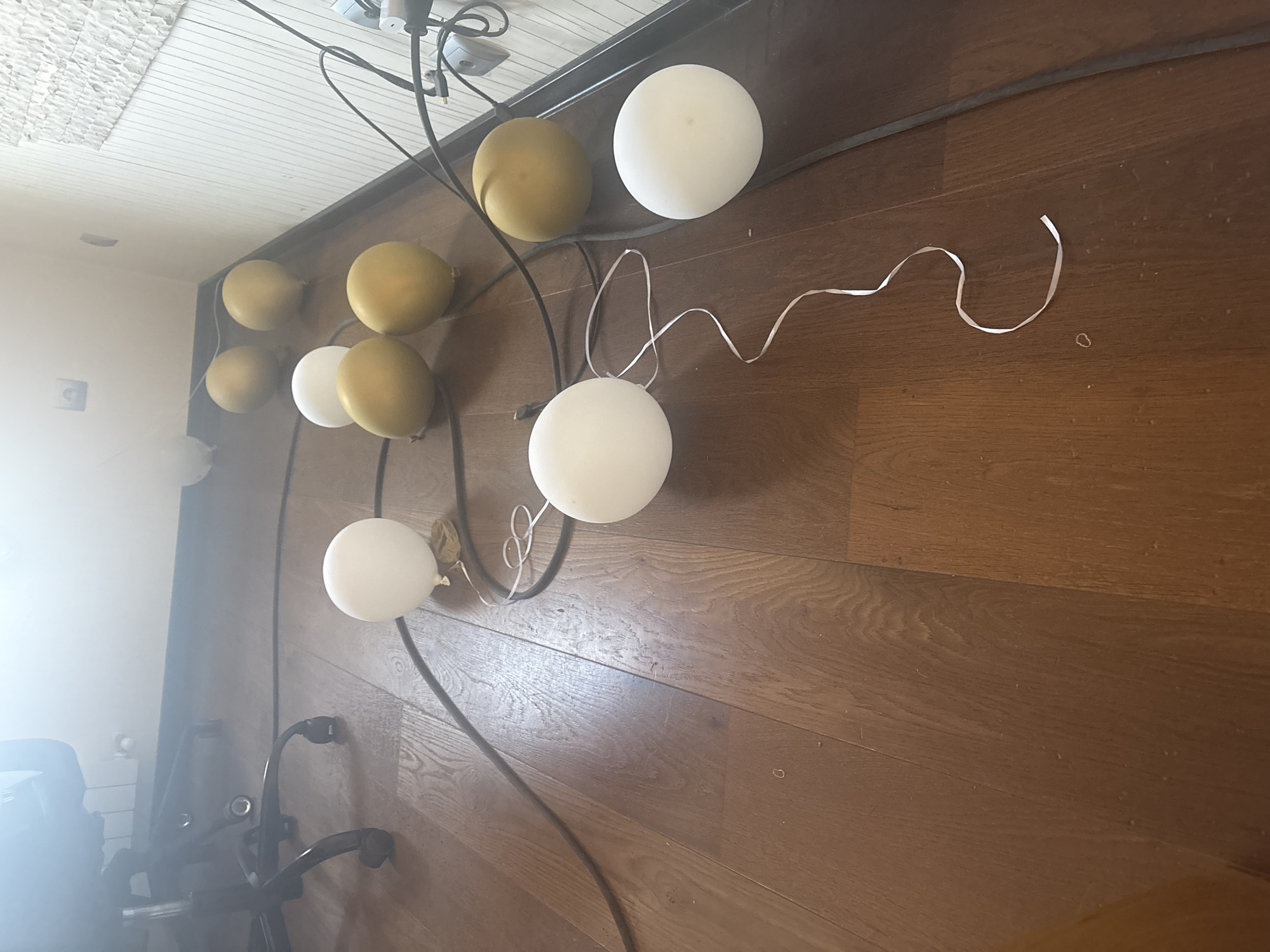 The image displays a cluster of deflated balloons and electrical cords on a wooden floor. The balloons are of two colors: white and gold. Some balloons are lying flat, while others appear partially inflated. The electrical cords are black and snake across the floor, connecting to what appear to be power outlets or fixtures on the wall in the background. A portion of an office chair with wheels is visible in the upper left corner. The wall in the background is light-colored and textured. The lighting in the scene suggests an indoor environment, possibly a room in a residential or office building. The location is identified as Orekhovka, Russia.