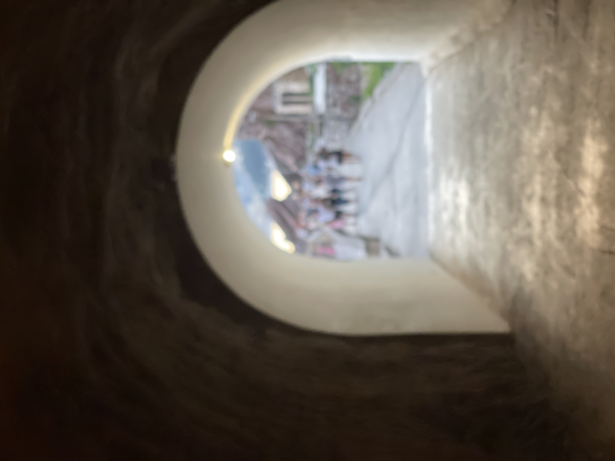 The image shows a view through a curved opening in a stone wall, looking out onto a scene in Rafailovići, Montenegro. The opening has a light-colored, smooth interior, and a small, bright light fixture is visible on its left edge.
Through the opening, a path or street is visible, lined with buildings. Several people are gathered on the path, some appearing to be seated. The background suggests an outdoor environment, with hints of greenery. The lighting indicates it is daytime, possibly late afternoon or early evening, given the soft, diffused light and the warm tones.
The texture of the stone walls within the opening is rough and dark, contrasting with the smooth interior of the archway and the bright, out-of-focus scene beyond. The overall impression is one of looking from a sheltered, possibly underground or interior space, out to a bustling area. The blurriness of the image obscures specific details about the people's activities, their clothing, or the architectural style of the buildings. No text is visible in the image.