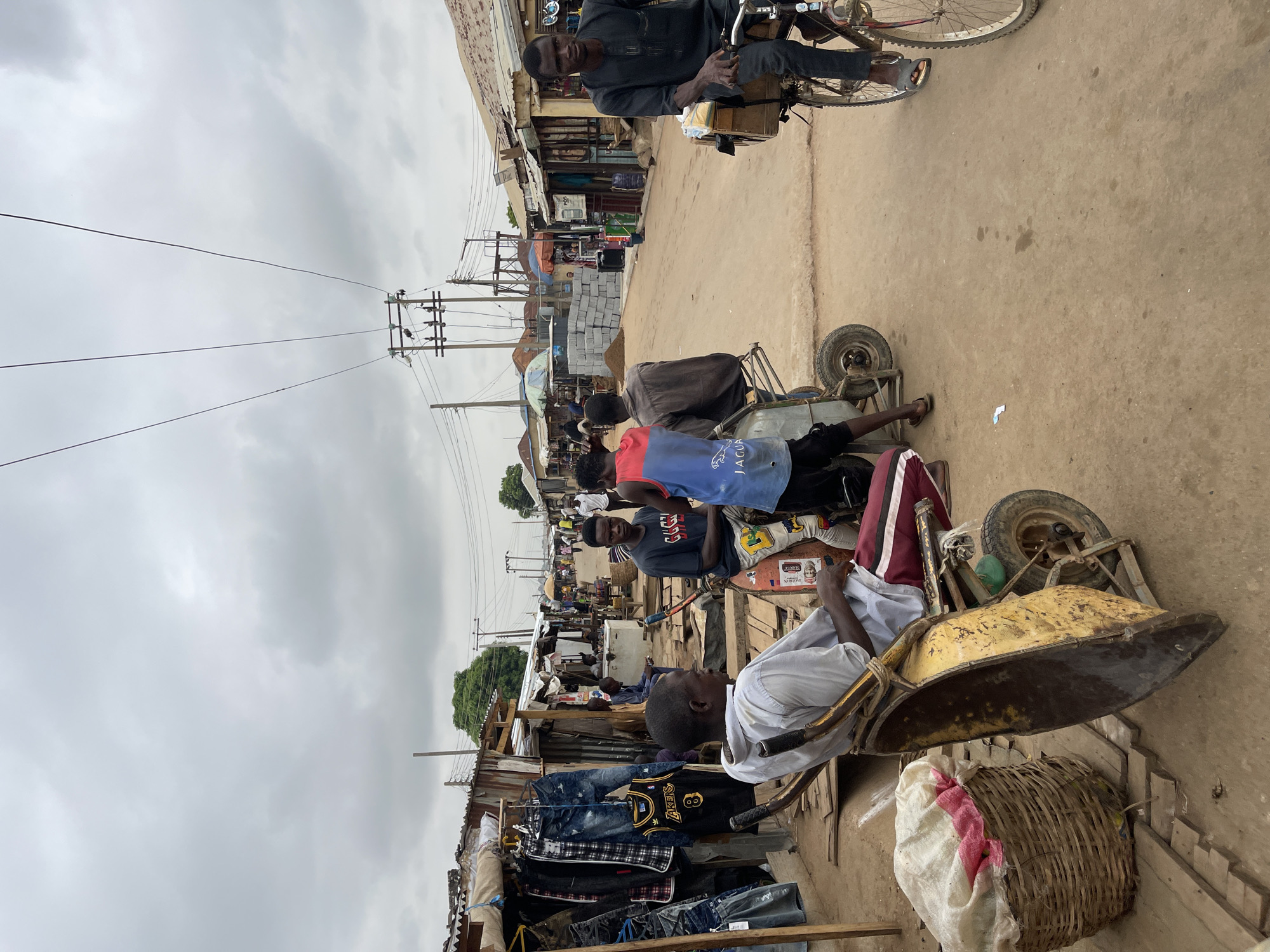 The image depicts a street scene in Kuchingoro, Nigeria. Several people are present, some of whom appear to be resting or waiting with wheelbarrows. Clothing is displayed for sale along the side of the road. Power lines run overhead. A man rides a bicycle in the distance. The street surface is dirt or sand. The sky is overcast.