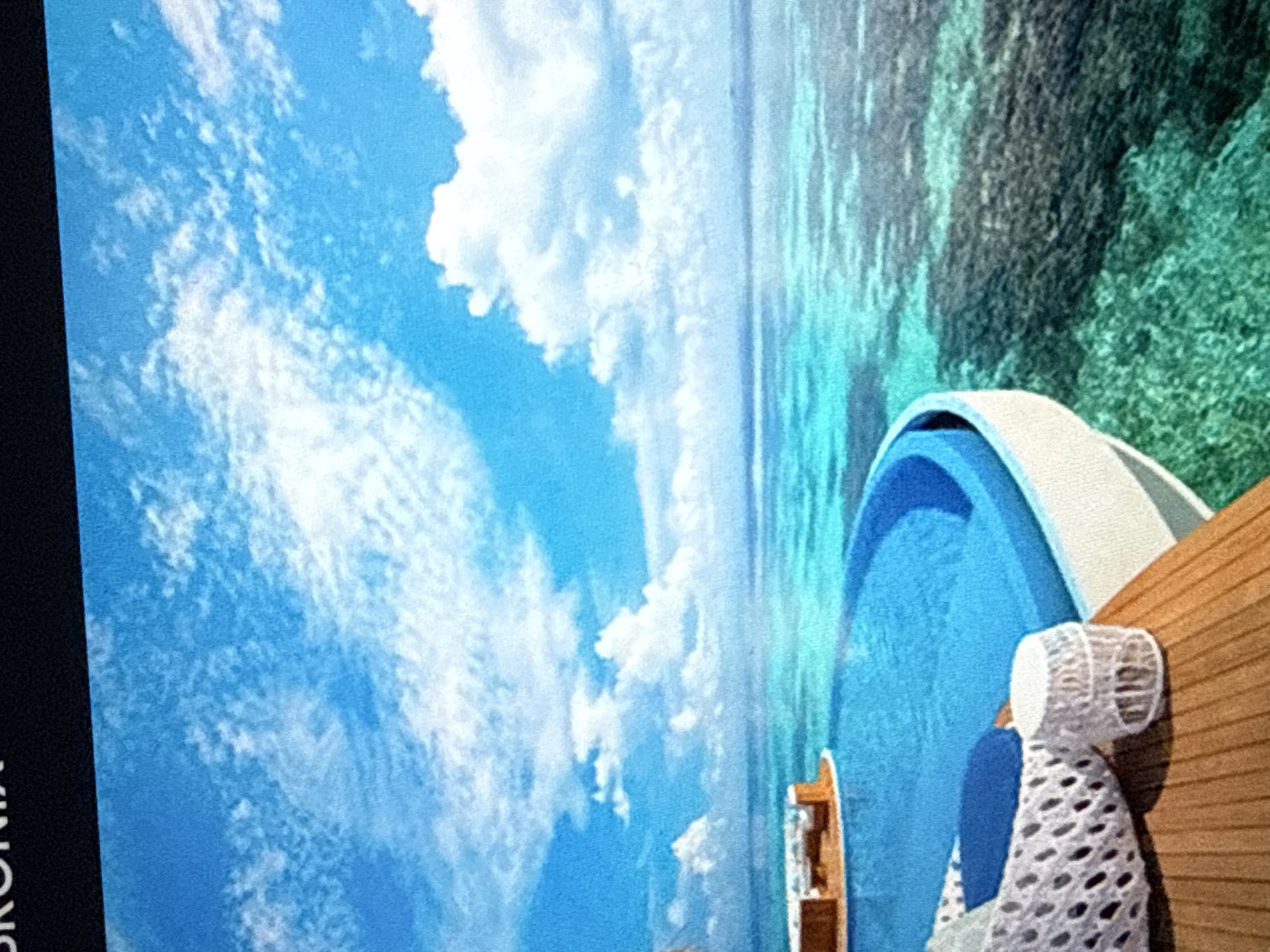 A digitally rendered image shows a resort with an infinity pool overlooking a tropical ocean. The pool is a vibrant blue, curving around the edge of a wooden deck. On the deck, there is white woven chair with blue cushions and a small, white cylindrical stool. In the distance, a circular structure with lounge furniture sits in the shallow, clear turquoise water. The ocean floor is visible, with dark coral formations and lighter sandy patches. The sky is a bright blue with scattered white clouds. The image appears to be a promotional rendering for a luxury resort.