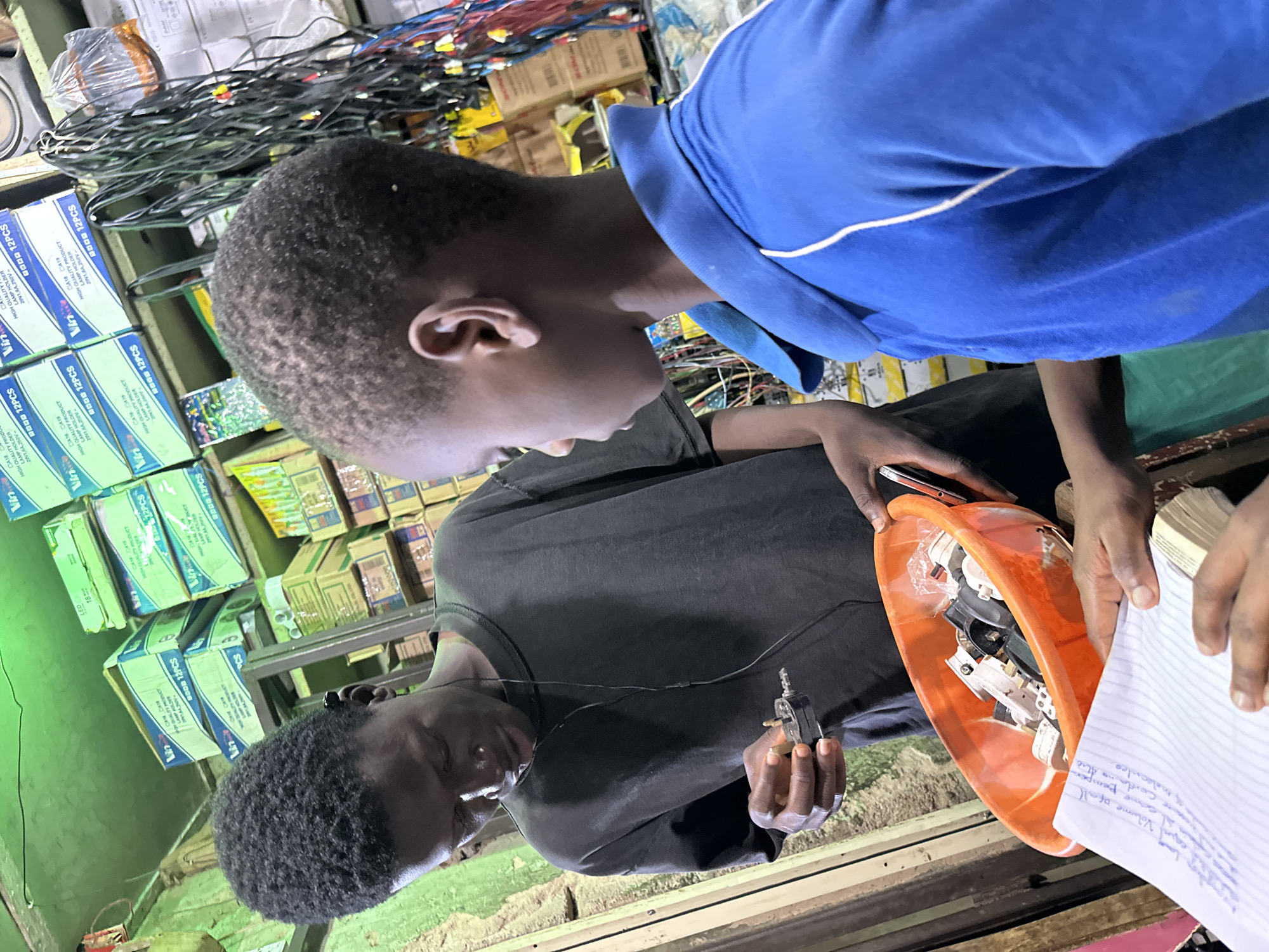 In the city of Narayi, Nigeria, two young individuals are engaged in an activity within a shop. The individual on the left, wearing a black t-shirt, is holding a small electrical component, possibly a plug or adapter, in his right hand and appears to be examining it. He has earbuds in his ears, suggesting he is listening to something. He is looking down at the component with focus. In front of him, an orange plastic bowl is filled with various electrical plugs and adapters.
The second individual, on the right, is wearing a blue polo shirt with white trim. He is facing the first individual and looking towards the bowl of electrical components. His left arm is resting on the bowl. In the foreground, a piece of paper with handwritten text, likely a list or notes, is visible, held by unseen hands.
The background of the shop is cluttered with shelves stocked with boxes, many of which appear to contain electrical items such as light bulbs and wiring, indicated by labels like "LED" and "12PCS." Hanging electrical wires and cables are also prominent in the upper right portion of the image. The lighting in the shop appears to be artificial, with a green tint coming from overhead. The overall environment suggests a retail setting for electrical goods.