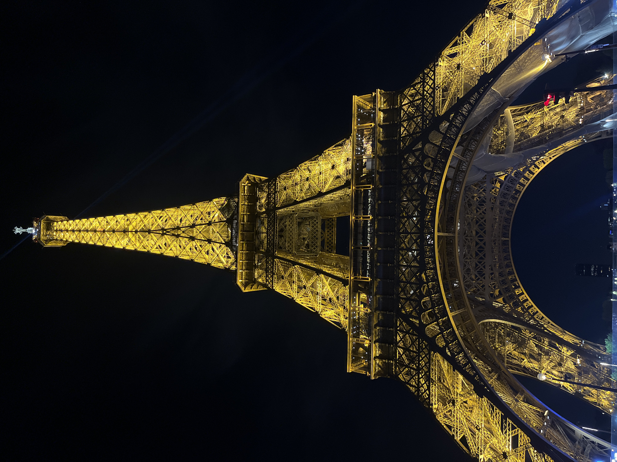 The image shows the Eiffel Tower in Paris, France, at night. The tower is illuminated with golden lights against a dark sky. The structure's latticework is visible in detail. Some areas of the tower appear to be under construction or renovation, with scaffolding and protective coverings present. The photo is taken from a low angle, looking upwards.