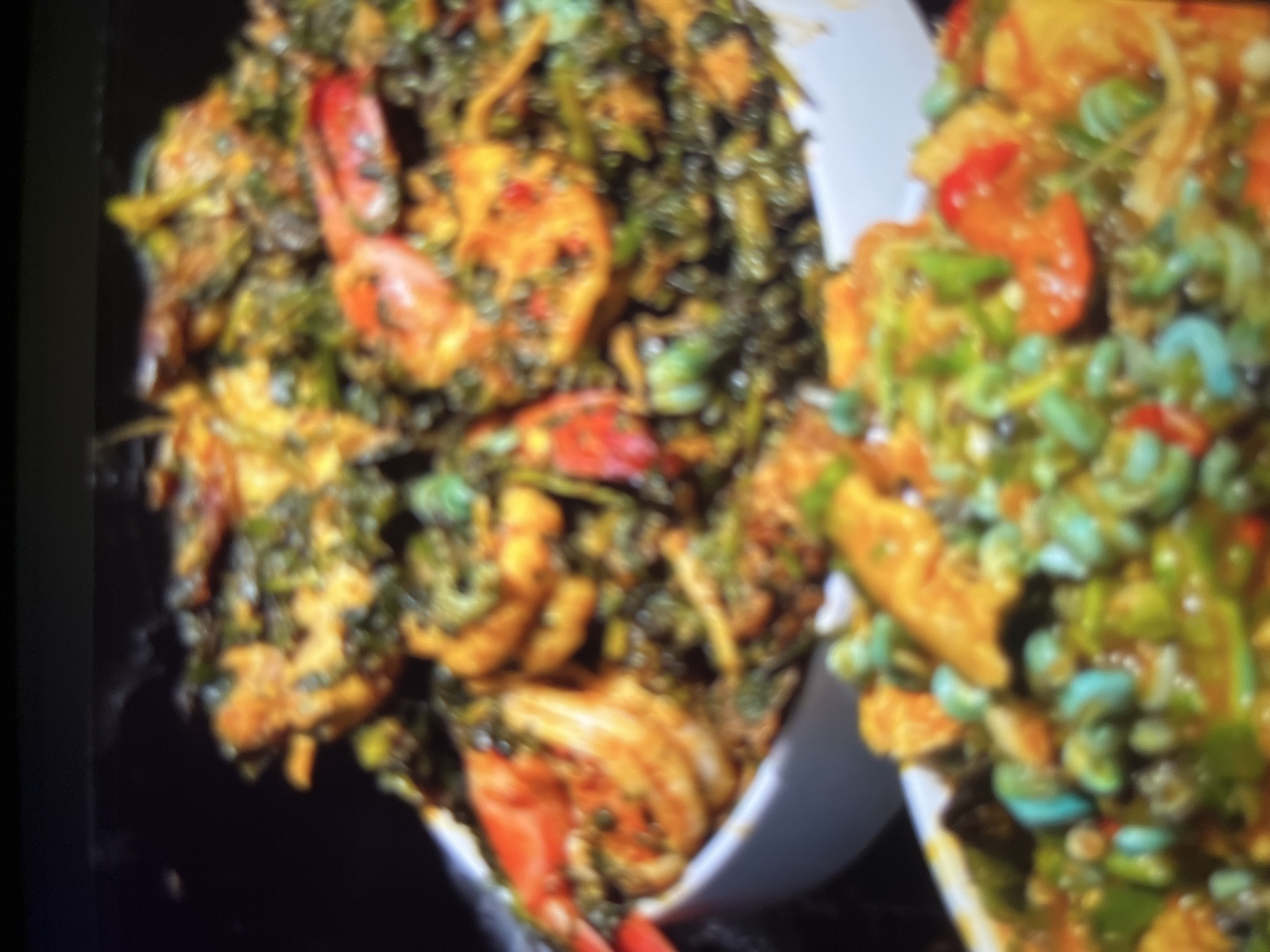 The image shows two bowls of food. The bowl in the foreground contains a dish with a green base, possibly a vegetable stew, with visible pieces of red pepper and what appear to be small, blue-green beans or peas. The bowl in the background is filled with a similar green stew, but it is dominated by large, pinkish-orange shrimp. The food is rich in color and texture, suggesting it is a flavorful and hearty meal. The lighting is dim, and the overall image quality is soft, making it difficult to discern fine details. There is no discernible information about the location in Abuja, Nigeria.