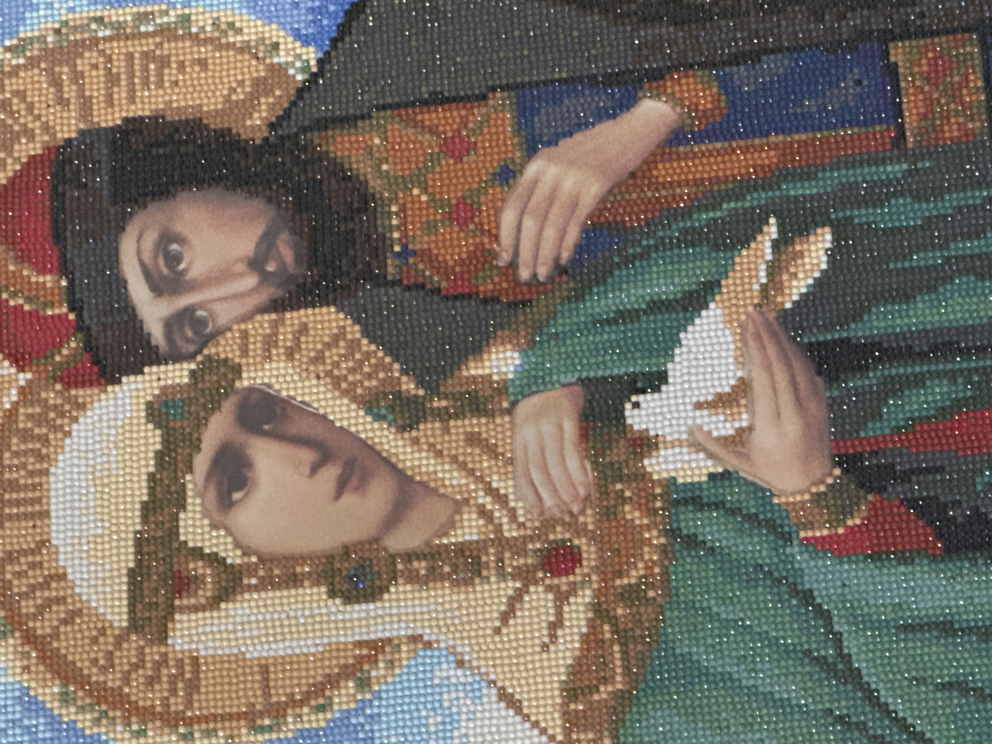 The image displays a religious artwork, likely an icon, rendered in the style of a diamond painting, characterized by its shimmering, bead-like texture. The artwork appears to be photographed rotated 90 degrees clockwise from its intended upright orientation.
The scene features two haloed figures. The figure positioned on the left side of the frame (which would be the upper figure in an upright viewing) is a male, depicted with dark hair, a mustache, and a short beard. His eyes are wide and dark, gazing directly outwards with an intense expression. He wears a dark outer garment over a richly patterned inner tunic, featuring intricate designs in gold, orange, and red. His golden halo is simple. His right hand is visible, resting gently on his clothing.
The figure on the right side of the frame (which would be the lower figure in an upright viewing) is female, portrayed with a serene and contemplative expression, her gaze directed softly downwards. She is adorned with a more elaborate golden halo, accented with red and green jewel-like details. She wears a flowing green robe over a lighter garment. Her hands are clasped together, holding a white, bird-like object, most likely a dove, with its wings slightly spread.
The background is dark and indistinct, with subtle hints of blue visible at the top and bottom edges of the frame. The entire composition is made up of countless small, glittering beads, which create a unique texture and a sparkling effect, particularly noticeable on the halos and the patterned garments. No text is visible within the image. The artwork conveys a sense of spiritual reverence and peace.