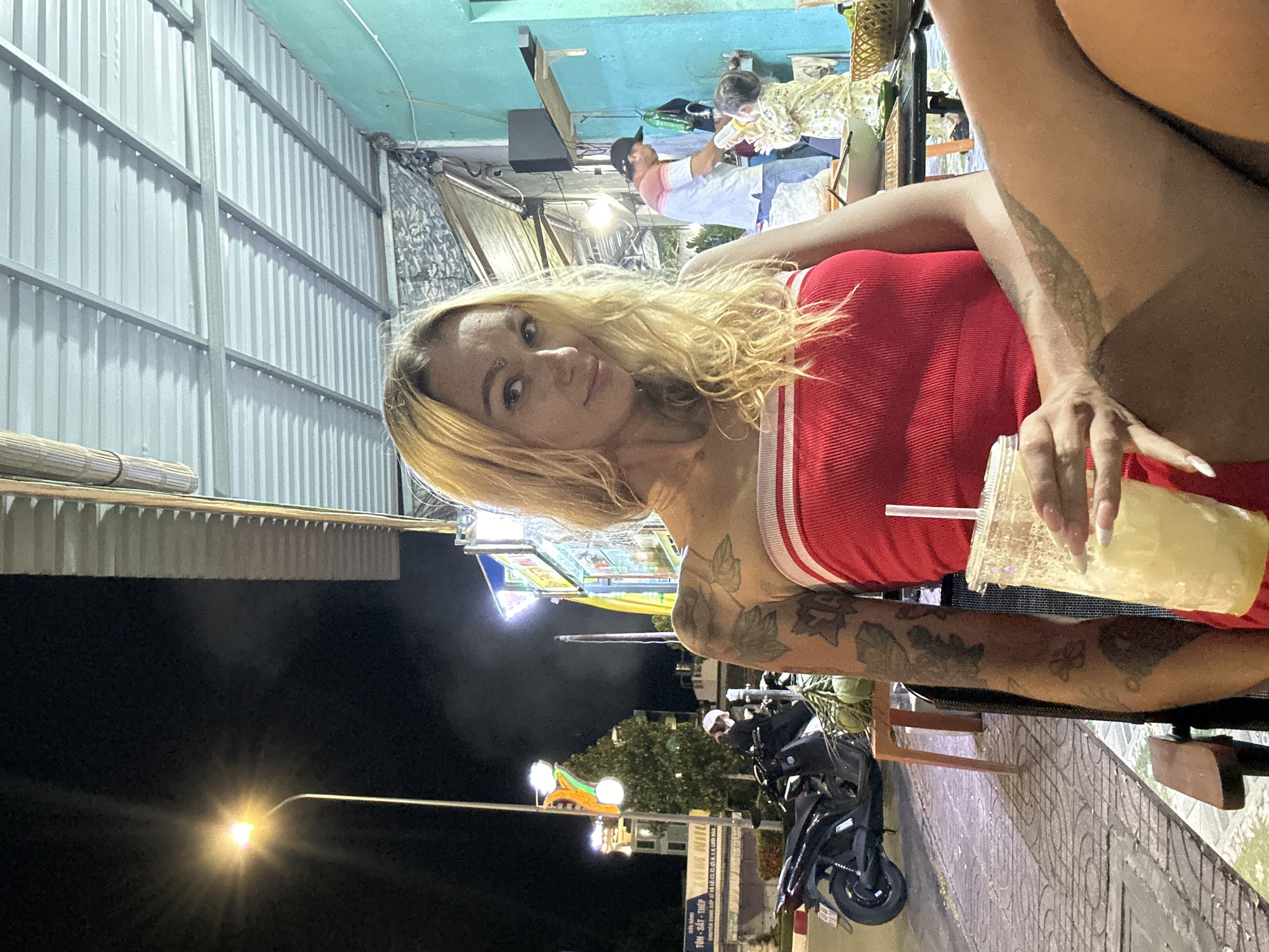 A woman with blonde hair and tattoos on her arm sits at a table outside at night in Phu Thuy Ward, Vietnam. She is holding a clear plastic cup with a straw. She is wearing a red strapless top. In the background, a man is standing and appears to be interacting with a child. There is a motorcycle parked nearby. The scene is illuminated by streetlights and lights from buildings.
