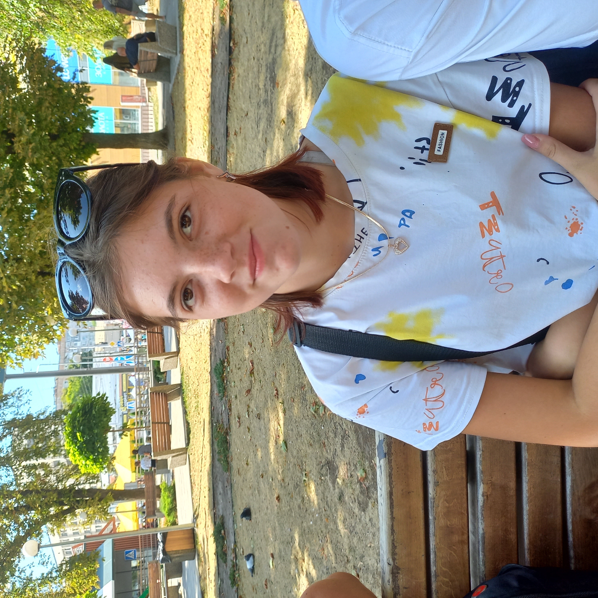 A young woman with brown hair and freckles, wearing a white t-shirt with colorful graffiti-style print, a gold necklace with a heart pendant, and sunglasses on her head, is sitting on a wooden bench in a park. The background shows trees with green foliage, buildings, and other people sitting on benches. The scene is brightly lit by sunlight, suggesting it is daytime. The location is the central park of Kropyvnytskyi, Ukraine.