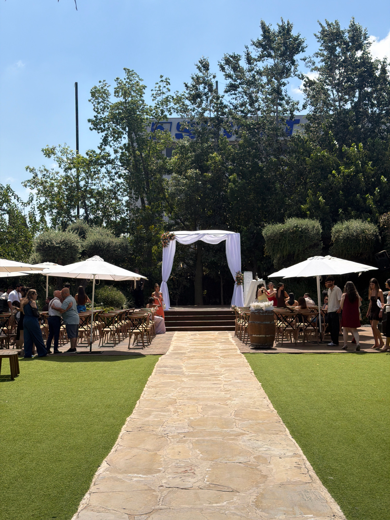 The image captures an outdoor wedding ceremony venue in Kadima, Israel. A stone pathway leads towards a decorated wedding arch draped with white fabric, situated on a raised wooden platform. Rows of empty wooden chairs are arranged on either side of the pathway, facing the arch, some covered by large white umbrellas. In the background, lush green trees and bushes surround the area, with a building featuring blue signage visible above the trees. Several people are scattered throughout the scene, some standing in groups, others appearing to be guests or wedding participants. The ground is a combination of artificial turf and the stone pathway. The sky above is clear and blue.