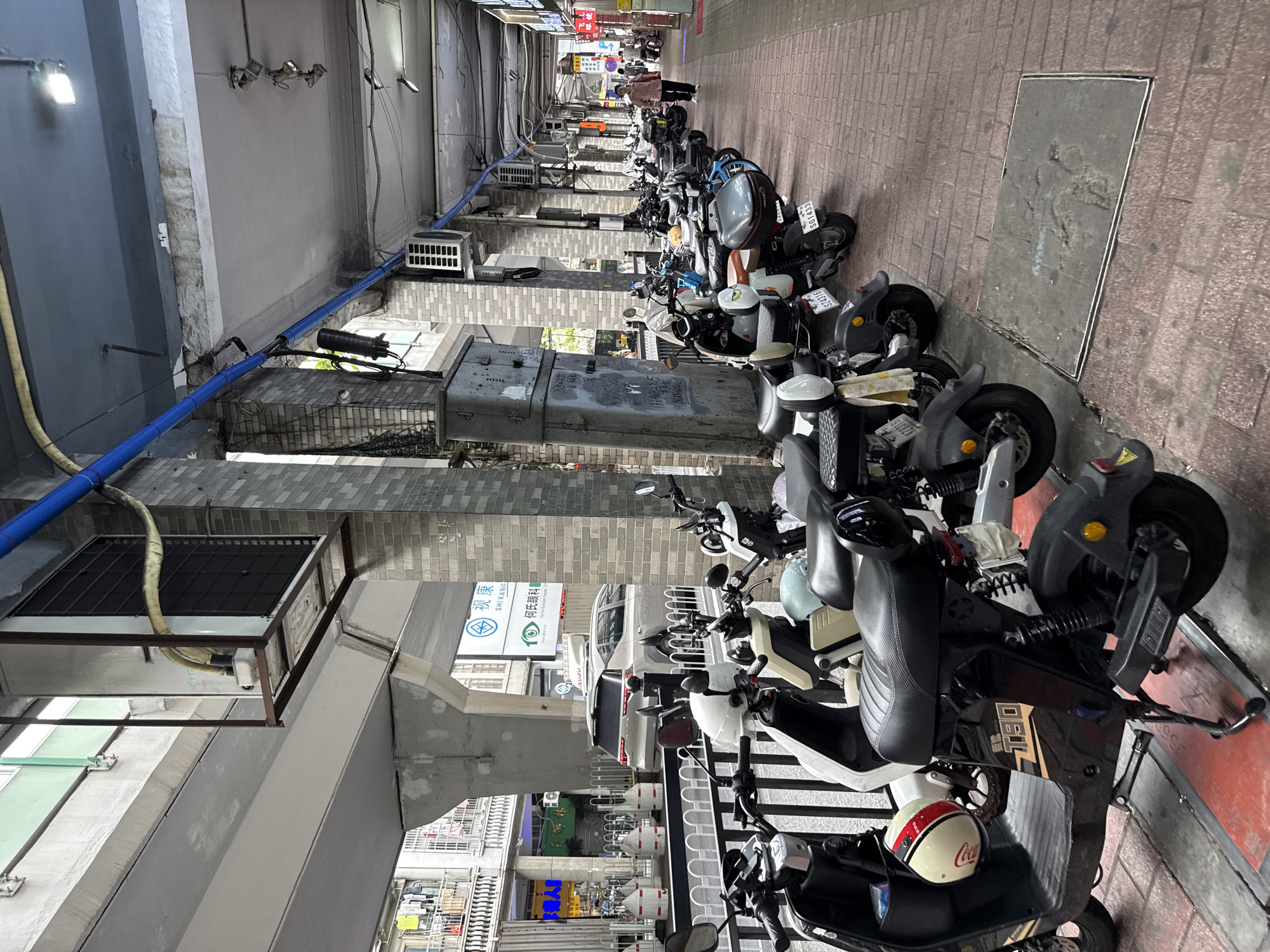 The image depicts a street scene in Yuexiu District, China, characterized by a row of parked electric scooters and motorcycles lining a brick-paved walkway. An elevated structure, possibly part of a bridge or overpass, casts a shadow over the scene. The architecture includes pillars with a brick facade and exposed concrete. Several air conditioning units are visible mounted on the exterior walls and under the elevated structure. A light-colored SUV is parked in the background, behind a white fence. In the distance, a woman in a pink shirt walks along the walkway, and other pedestrians are present. The overall environment suggests a public urban space with a focus on transportation and commercial activity, as indicated by storefronts visible in the background.