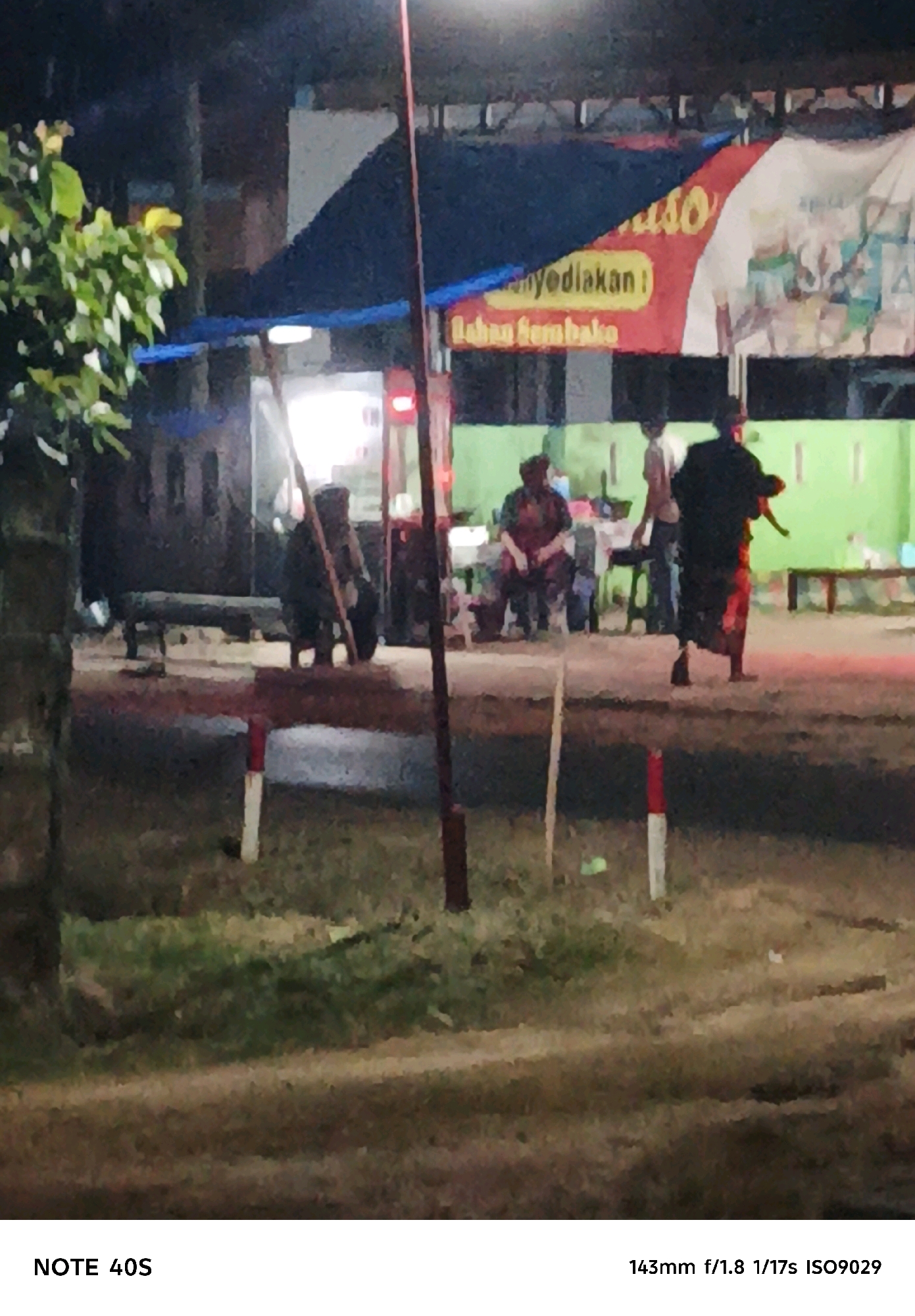 The image depicts an outdoor scene at night, likely in Adirejo, Indonesia. In the foreground, a person is walking away from the camera, dressed in dark clothing and a patterned skirt. To the left, another person is seated, wearing patterned clothing. Further back, more individuals are visible, some seated and some standing. A banner with text, partially obscured by darkness and the image quality, is displayed in the background. The environment includes foliage on the left, a street or pathway, and some structures that appear to be stalls or buildings. Red and white markers are present in the foreground, suggesting a roadside or boundary. The overall lighting suggests artificial light sources, possibly from lamps and the banner. The camera settings indicate a focal length of 143mm, an aperture of f/1.8, and a shutter speed of 1/17th of a second, with an ISO of 9029.