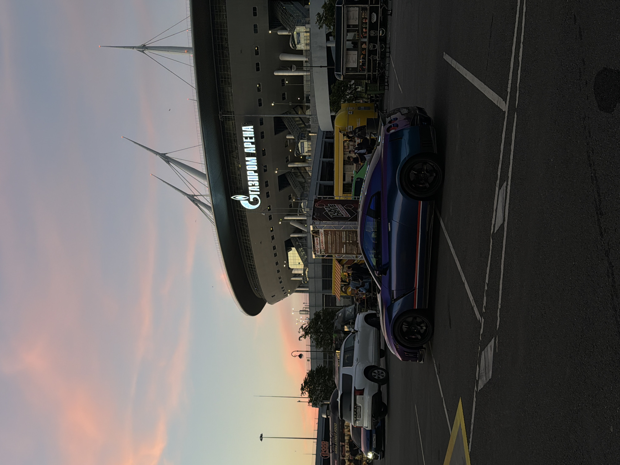 In an outdoor parking lot, a scene unfolds during a twilight hour with a soft, pink-hued sky. Dominating the background is the Gazprom Arena, a large, modern stadium with distinctive white spires reaching towards the sky, illuminated by the fading light. The stadium's name is prominently displayed in large, white letters.
In the foreground, a dark blue Nissan GT-R sports car is parked, its sleek design catching the low light. To its left, a white SUV and a dark blue sedan are also parked. Several food trucks are visible behind the parked cars, some with people gathered around them, suggesting an event or gathering. The ground is asphalt, marked with parking lines. The overall atmosphere is a blend of urban architecture and casual activity under a dramatic sky.