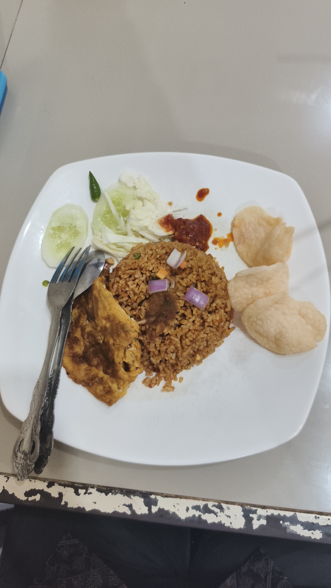 A white plate holds a serving of fried rice topped with diced red onion and a small pile of minced meat or spice paste. To the left of the rice is a folded omelet. The plate also contains cucumber slices, shredded cabbage, a single green chili, and a dollop of red chili sauce. On the right side of the plate are three light-colored prawn crackers. A fork and spoon rest on the left side of the plate, partially covering the omelet. The food is presented on a light-colored surface, possibly a table, with a textured, worn edge in the foreground. The lighting suggests an indoor setting, likely daytime. There is no visible text in the image. The scene depicts a meal, likely lunch or dinner, in what appears to be a casual dining environment. The location context is Lhokseumawe, Indonesia.
